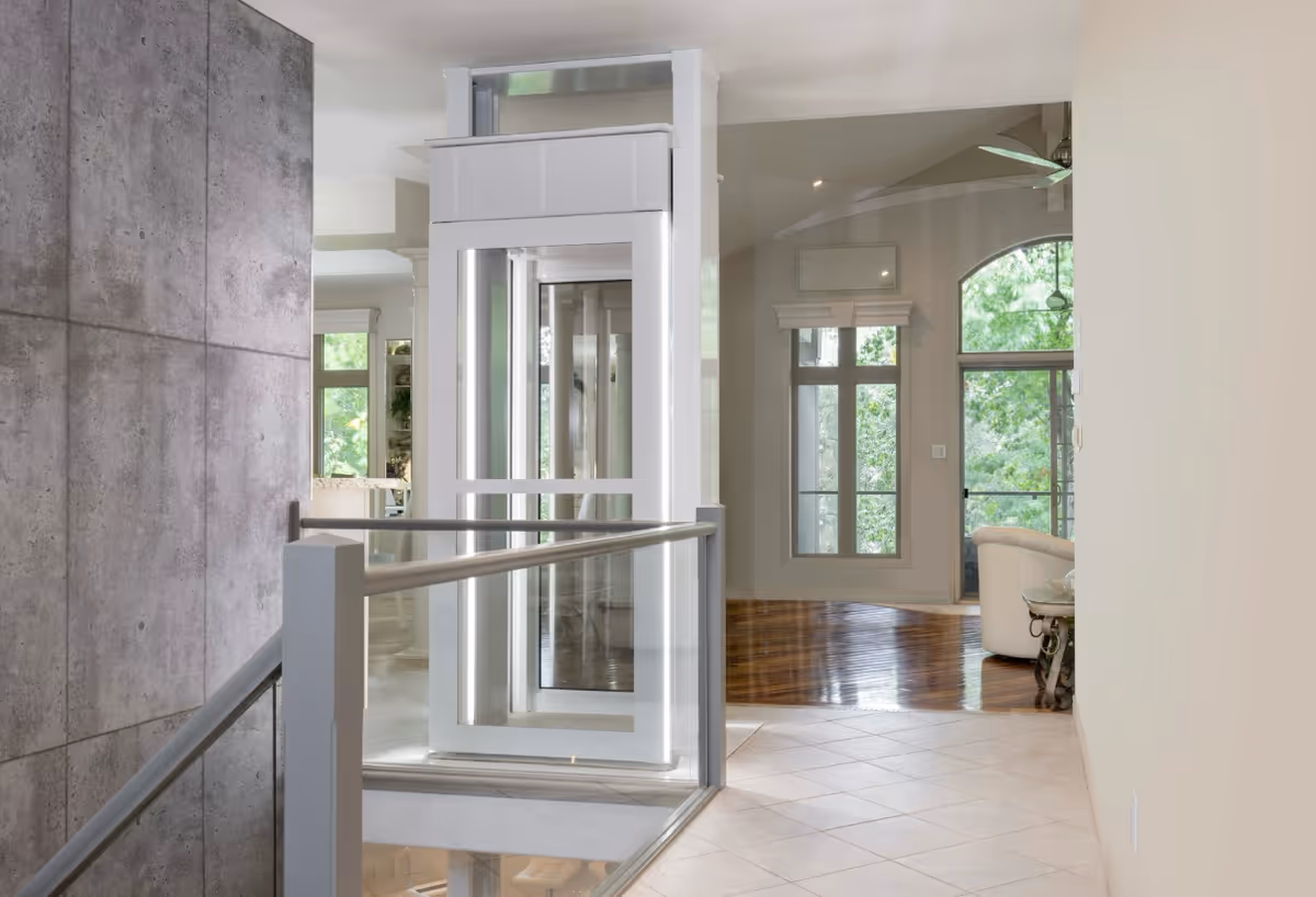 Modern interior with glass elevator, railing, tiled floor, and living area beyond.