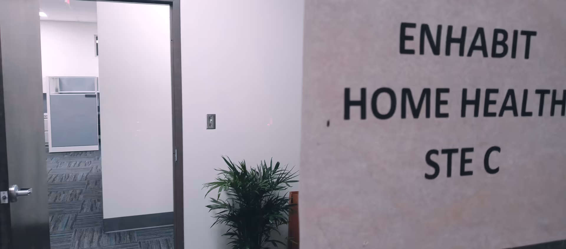 Corridor interior shows a doorway, a potted plant, and a wall sign reading Enhabit Home Health.