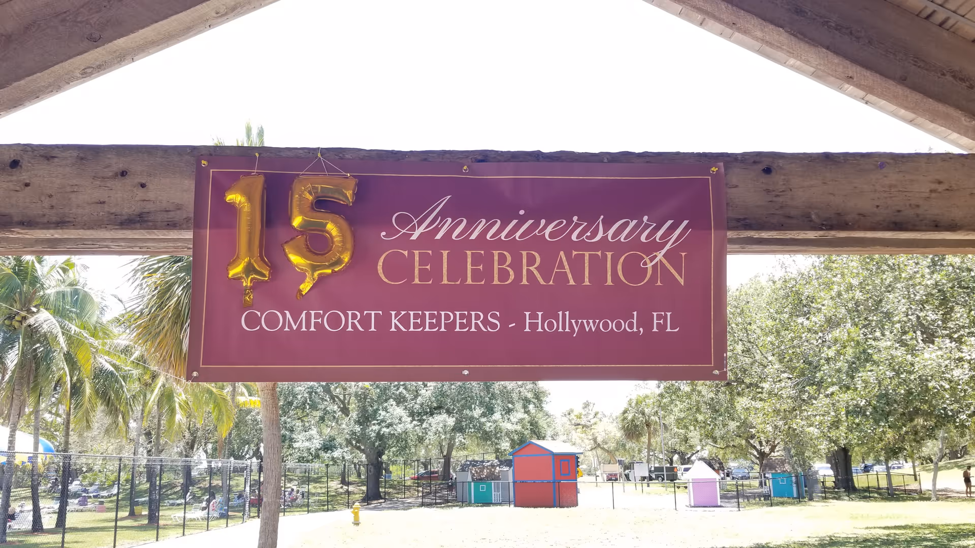 Purple 15th anniversary banner for Comfort Keepers Hollywood, FL hangs under a wooden outdoor pavilion.