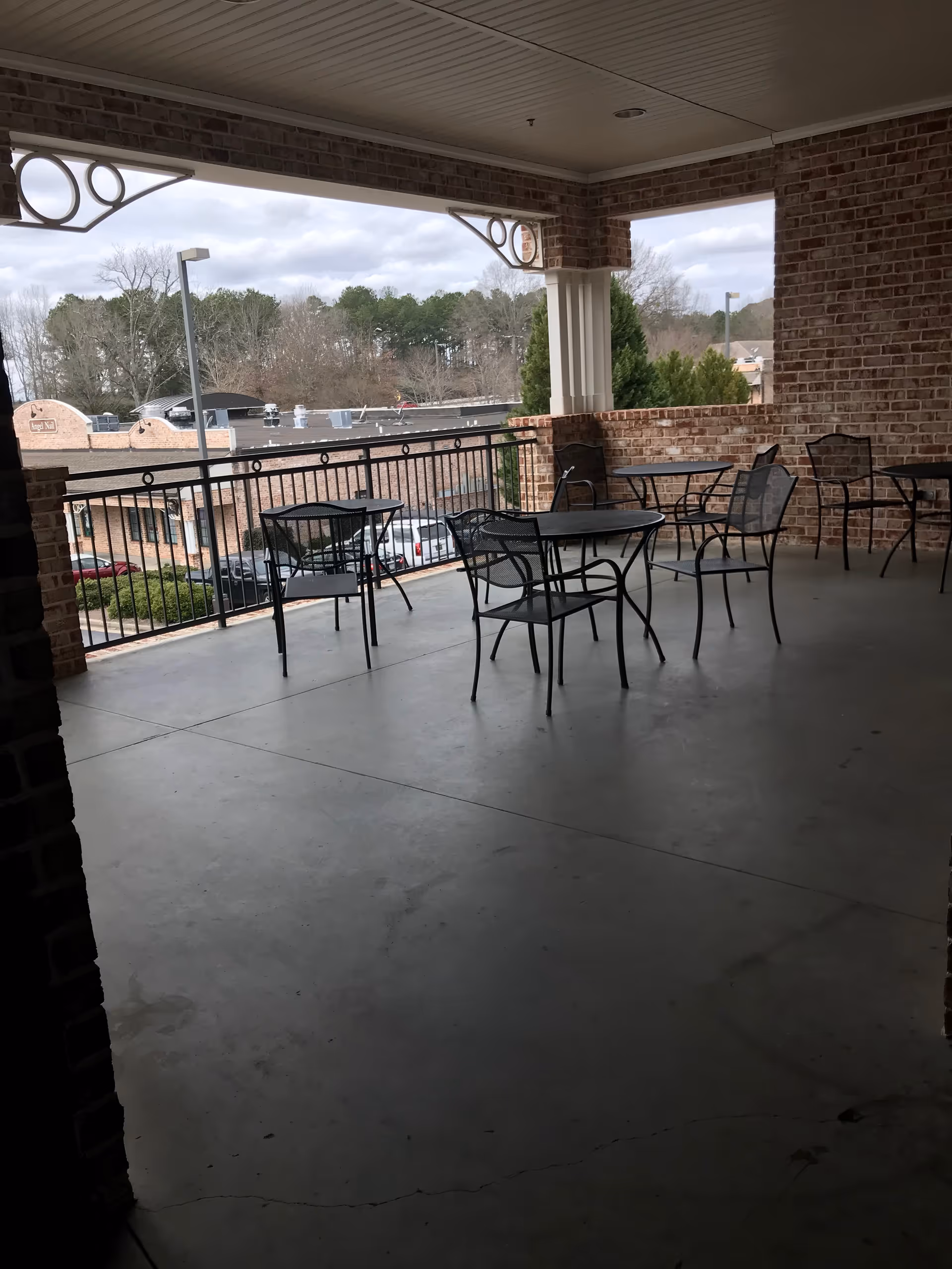 Covered outdoor patio with metal tables and chairs overlooking a parking lot and trees.
