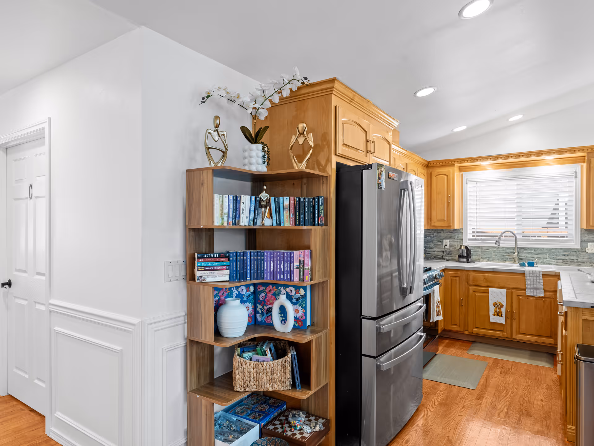 Bright kitchen with wooden cabinets, stainless fridge, and a tall bookshelf against a white wall.