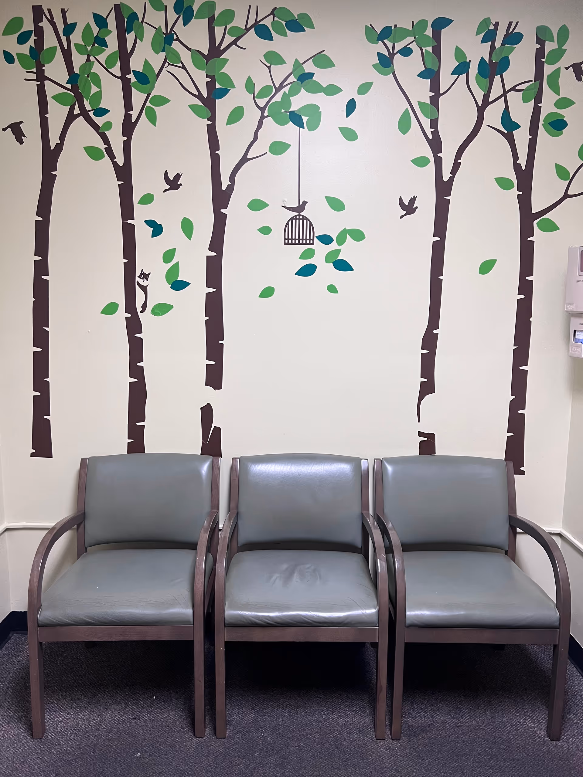 Three gray chairs sit against a wall featuring a tree mural indoors.