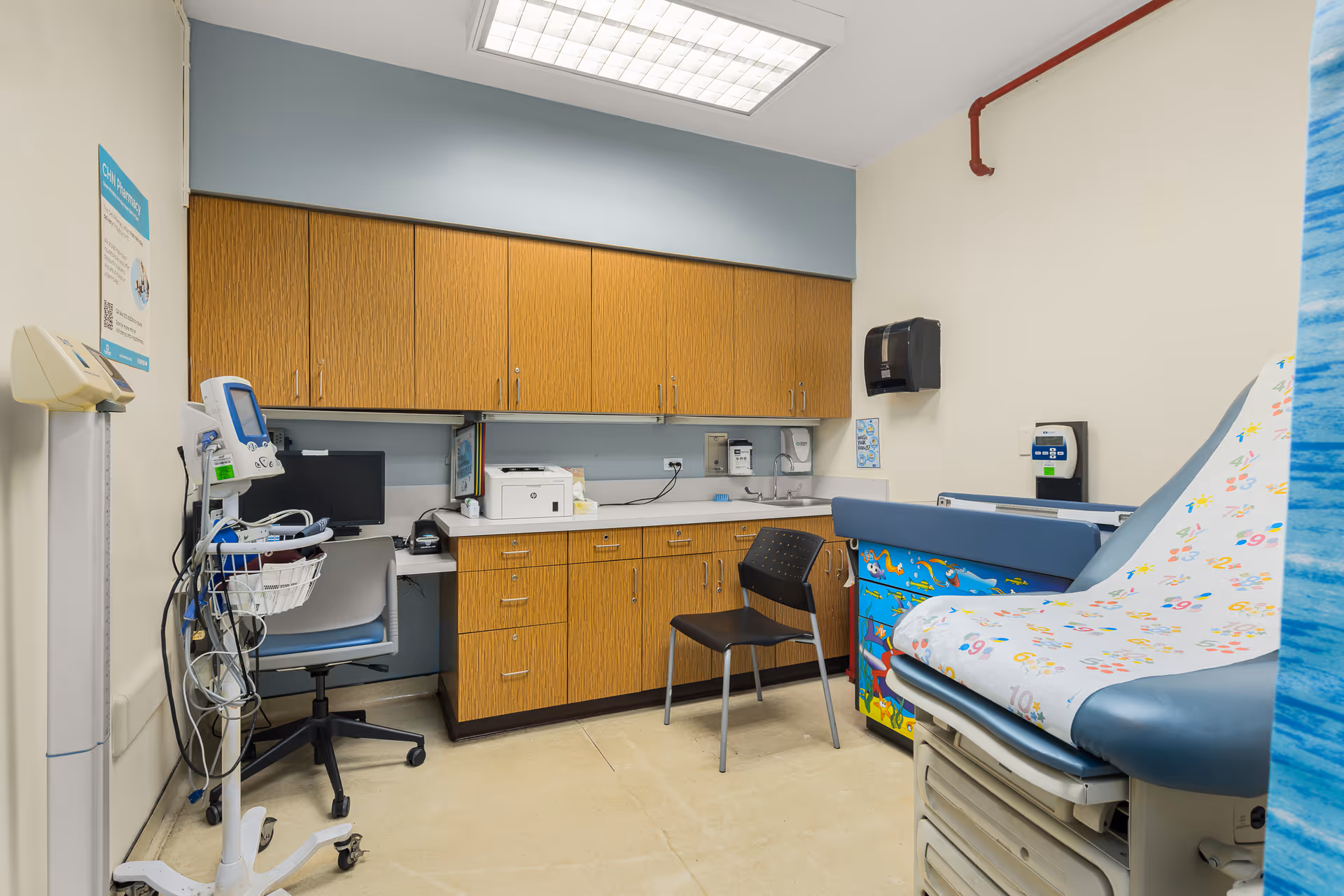 Clinical exam room with cabinets, computer, printer, and a colorful patient bed.