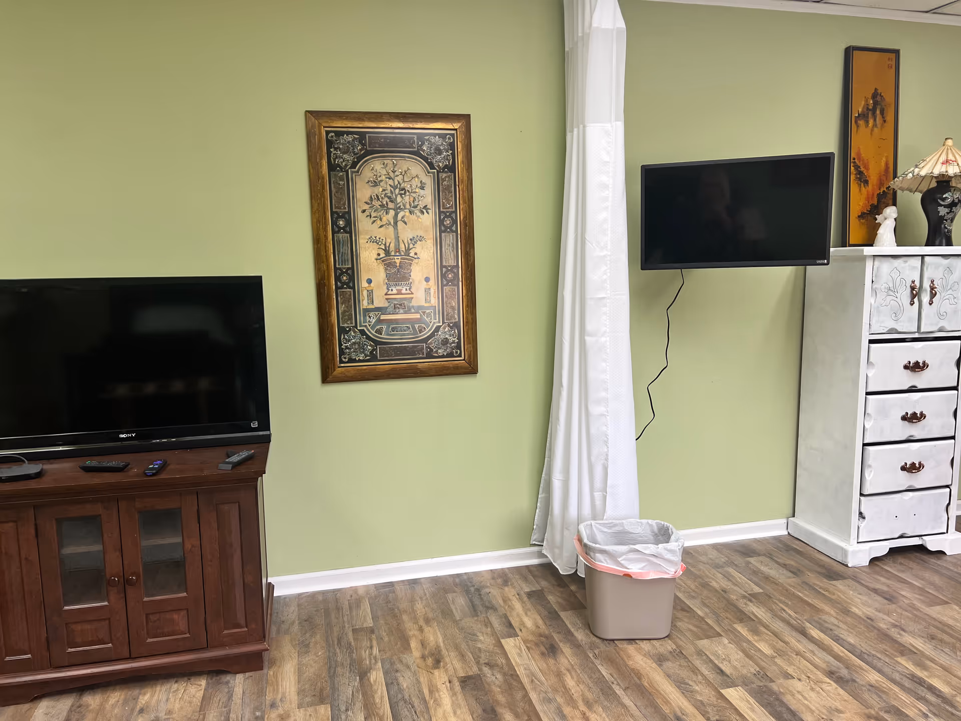 Green-walled living room with TVs, a cabinet, painting, curtain, and a trash can.