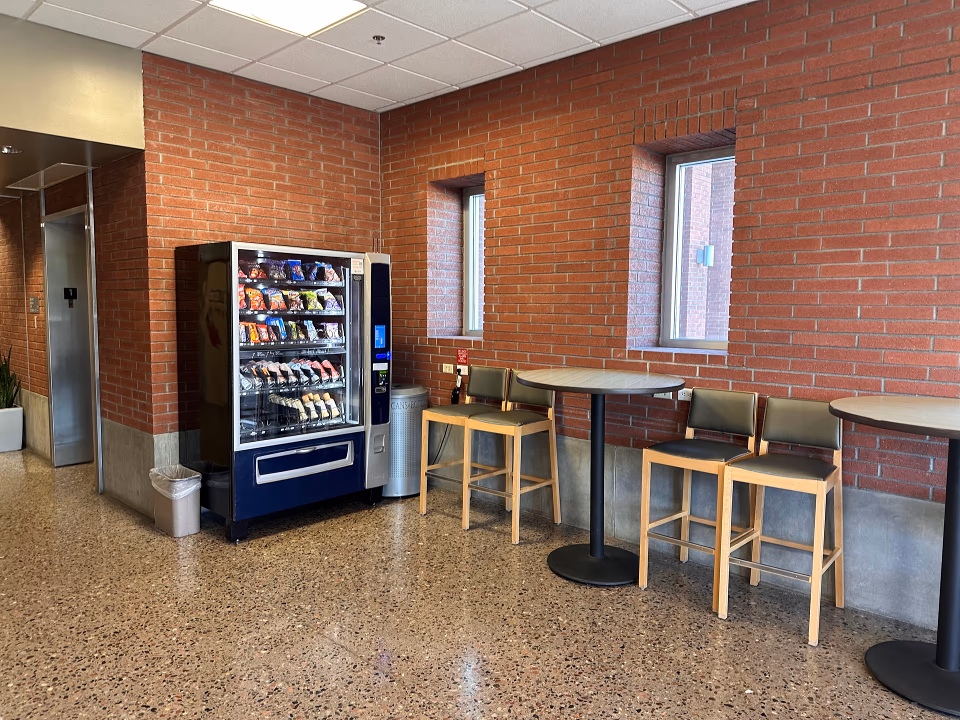 Vending machine with snack selection beside high tables along brick hallway.