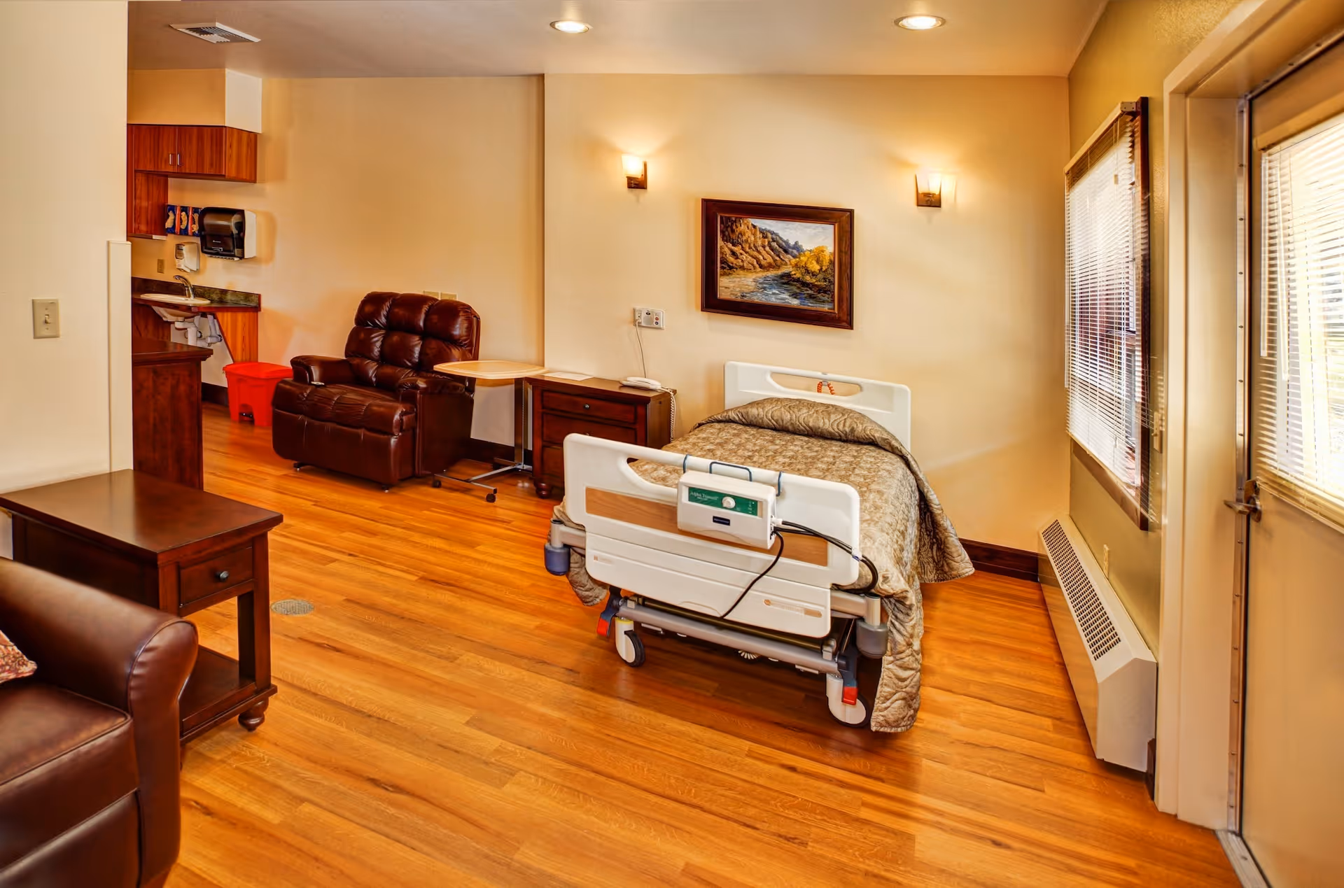 Hospice patient room with hospital bed, recliner, wood floors, and windowed door.