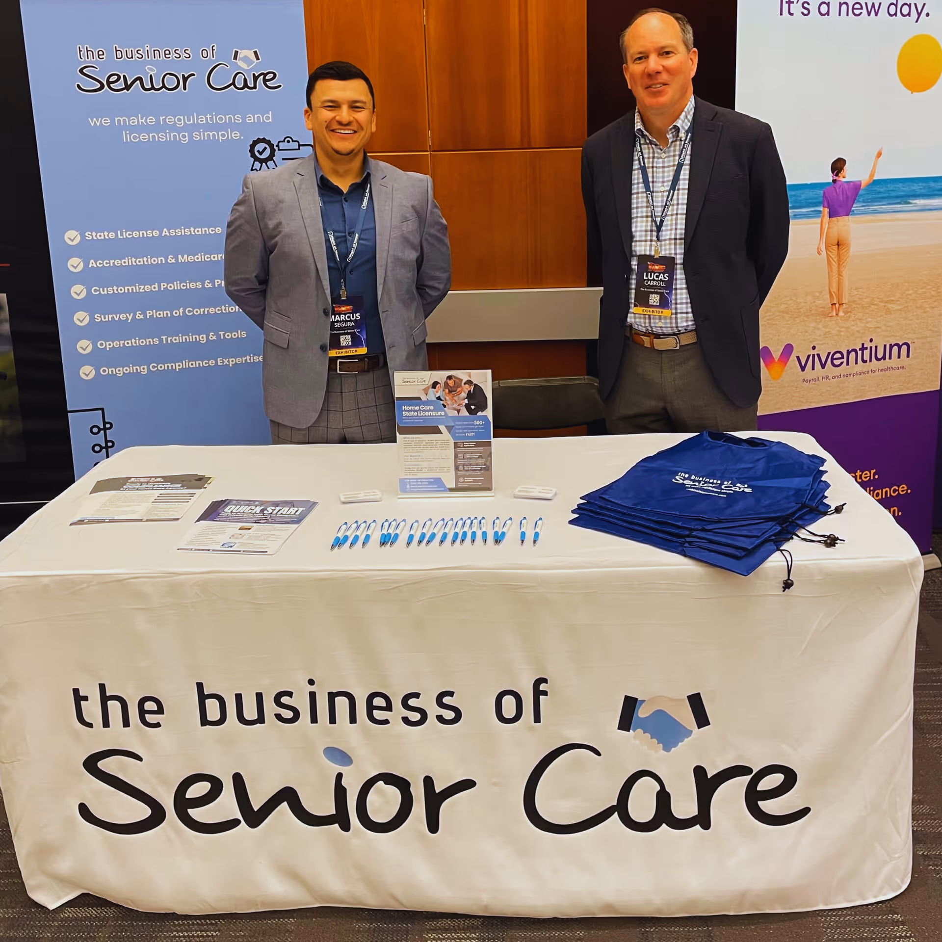 Two men stand beside a promotional booth for The Business of Senior Care.