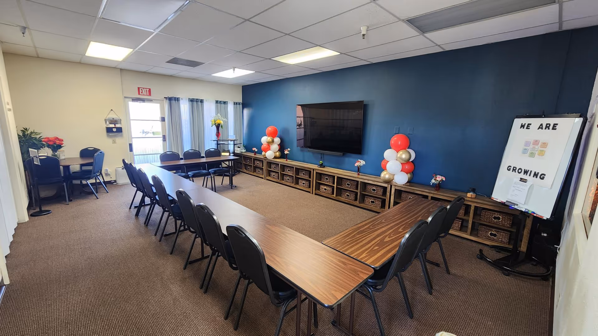 A large conference room with U-shaped tables, blue wall, TV, and balloons.
