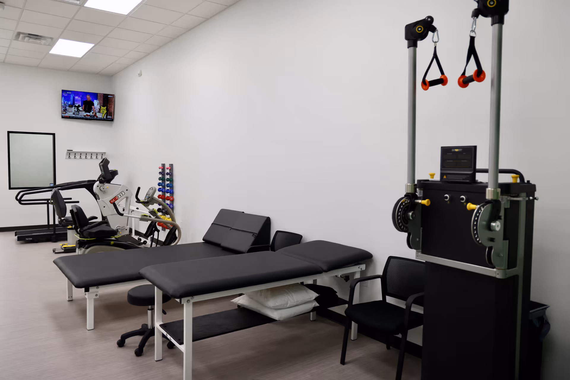 Interior rehab gym with treatment table, exercise machines, weights, and a wall-mounted TV.
