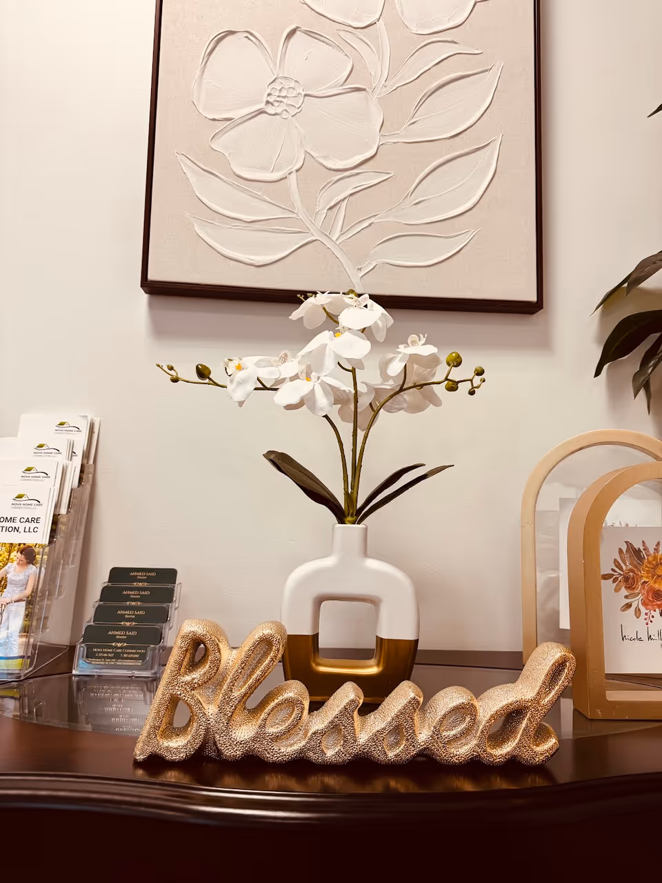 White orchid vase on a wooden table with framed art and a glittery 'Blessed' sign.