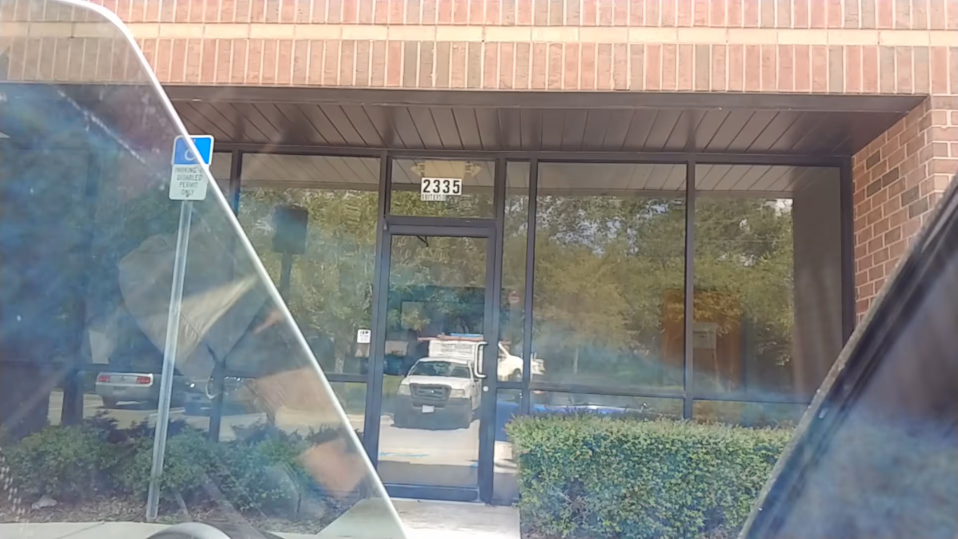 Brick building entrance with glass doors, hedges, and a parking lot reflected in glass.