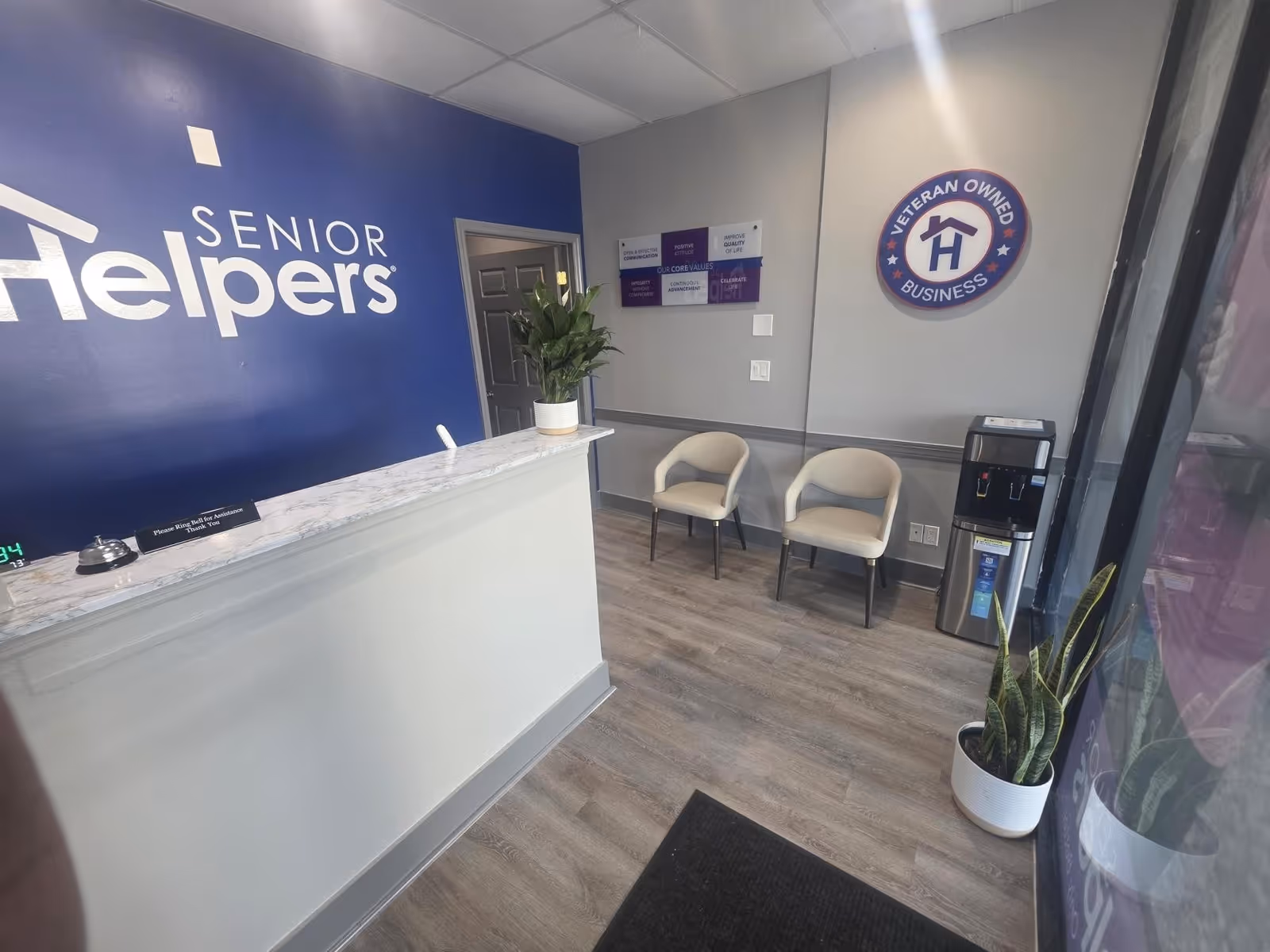 Reception area of Senior Helpers with blue wall, marble counter, chairs, water cooler, and plant.