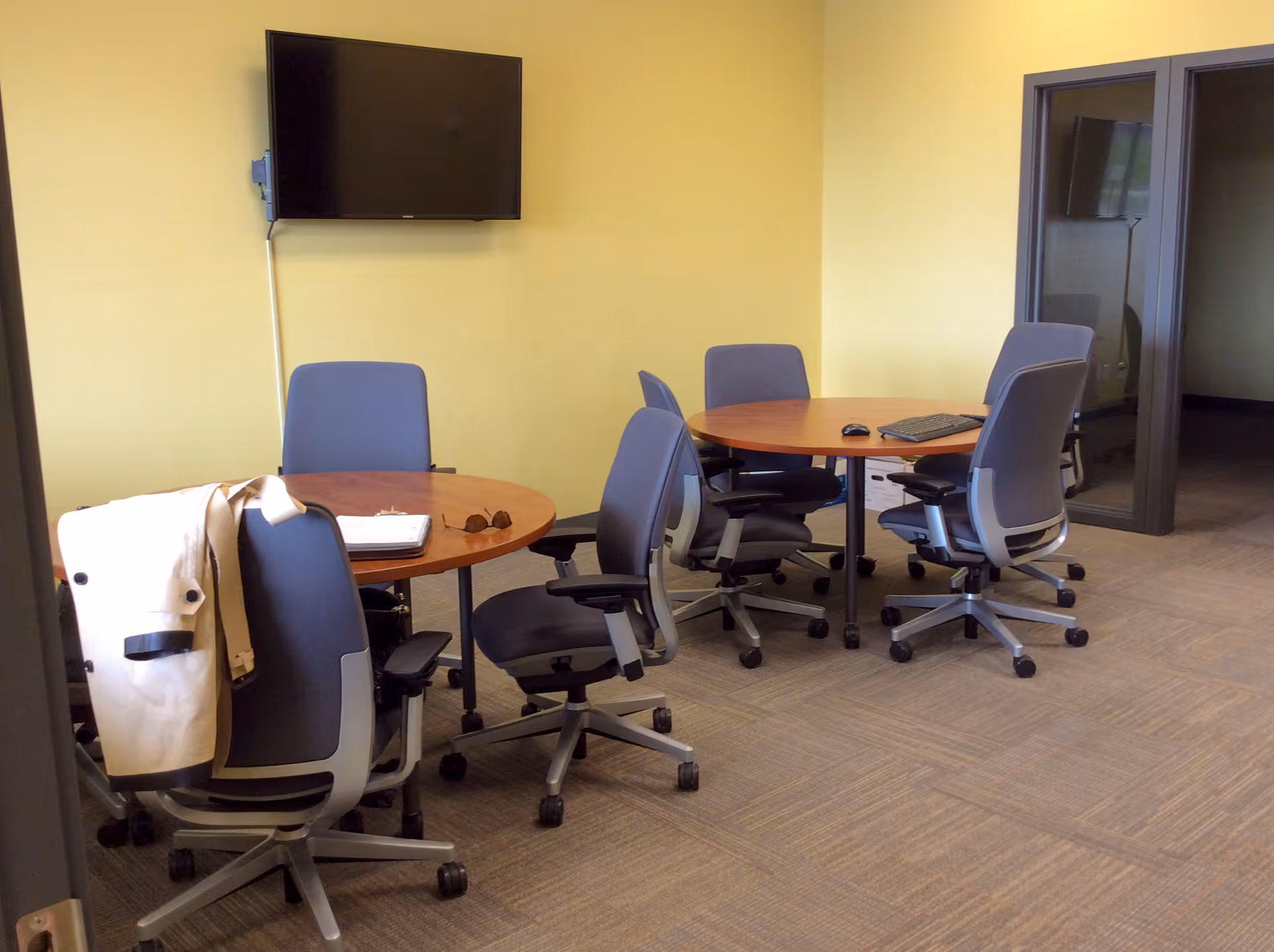 Yellow-walled conference room with round tables, blue chairs, and a wall-mounted TV.