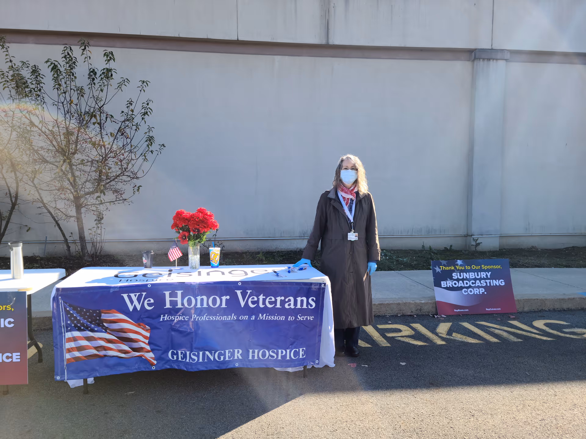 A person in a coat and mask stands beside a table with a veterans banner outdoors.