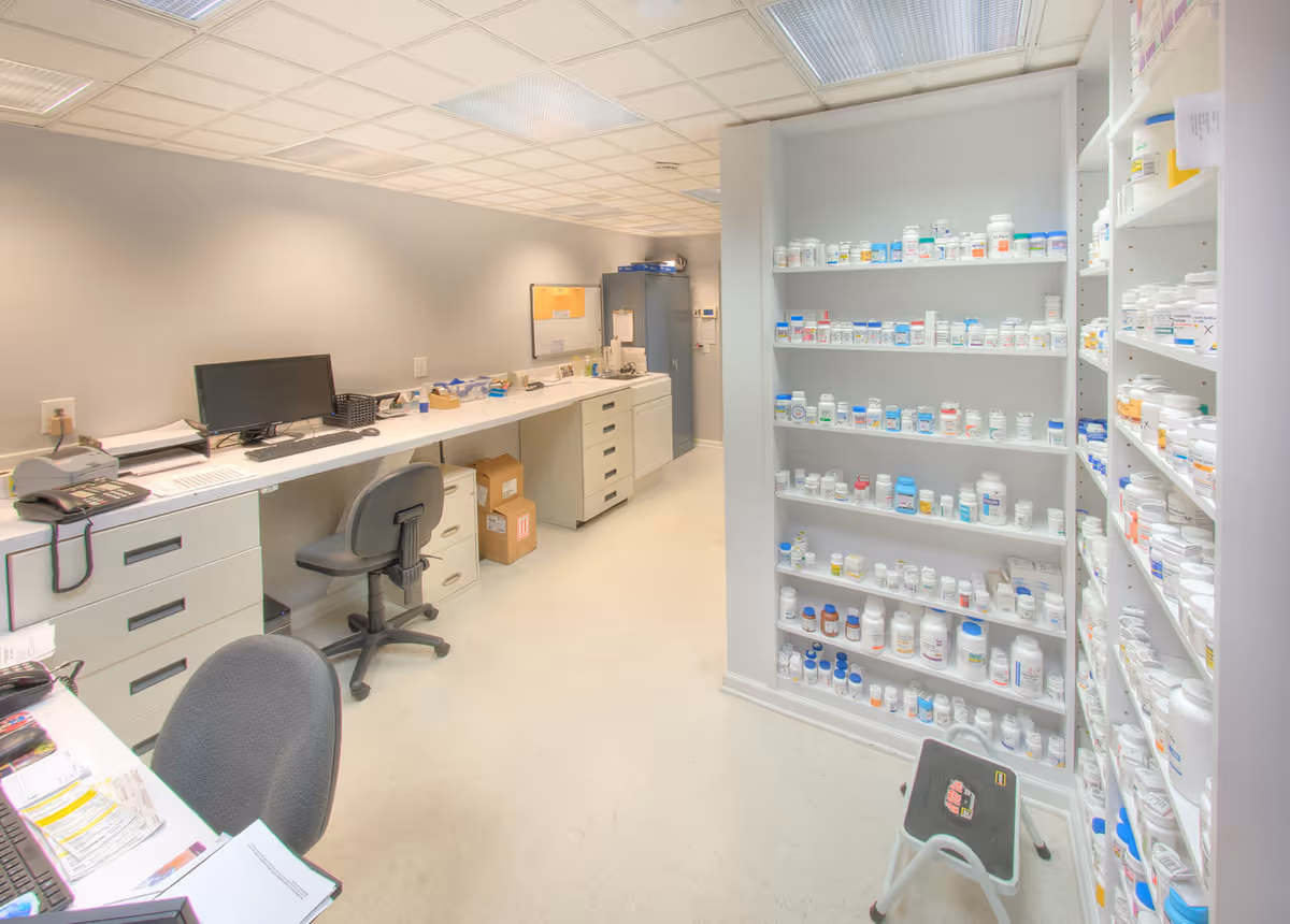 Long medical supply counter with computer, chair, and tall shelves stocked with bottles.
