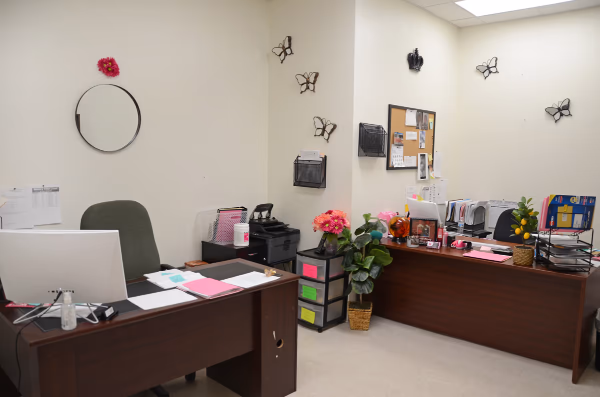 A tidy office reception area with two dark desks, chairs, bulletin board, and plants.