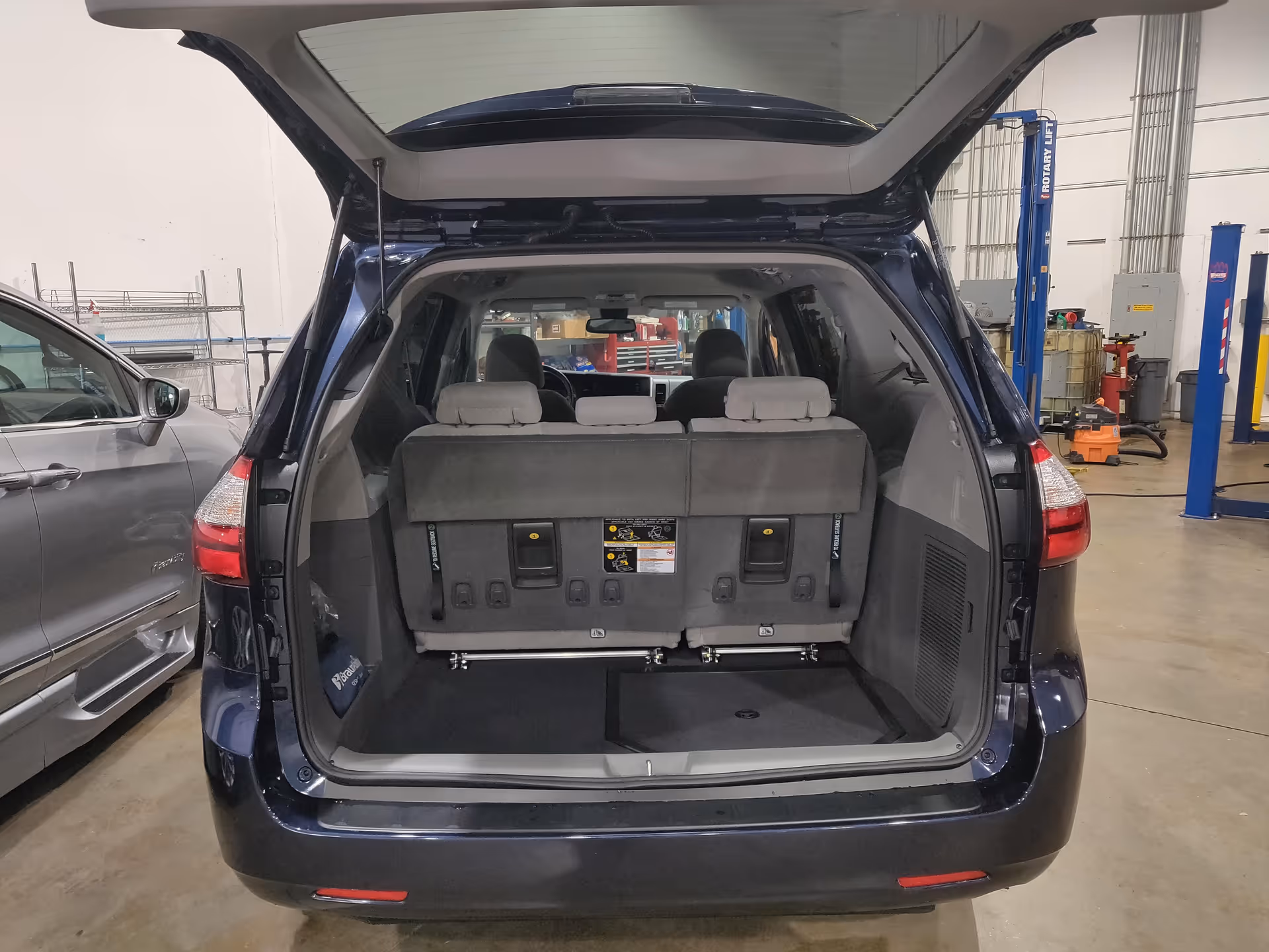 Open rear hatch of a dark blue minivan shows folded third-row seats in a workshop