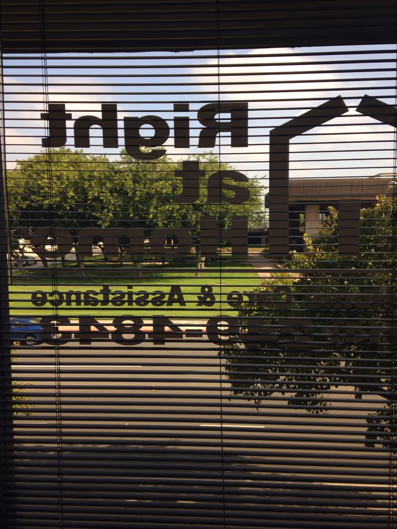 View through blinds of a Right at Home exterior with trees and a building