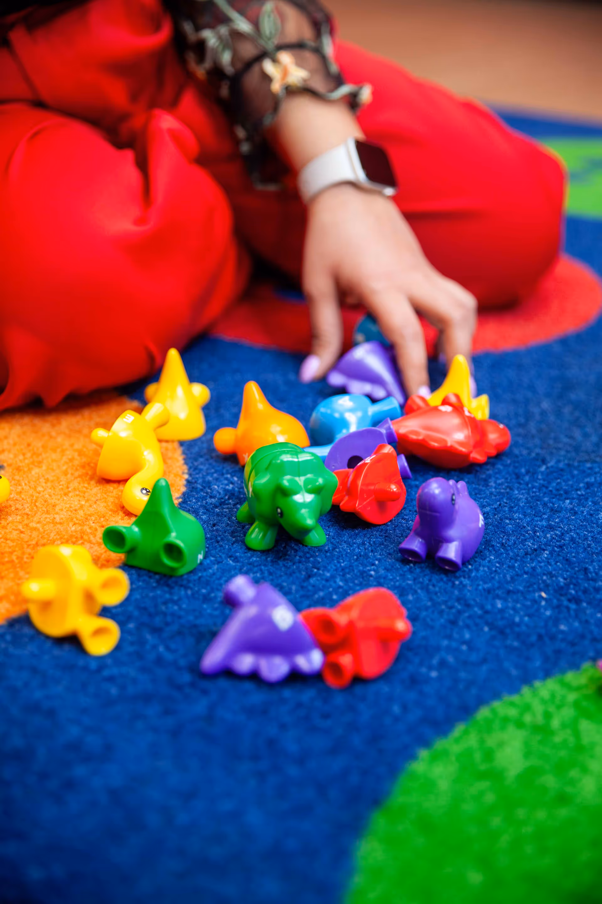 A person in red sits on a blue carpet, reaching for colorful animal-shaped toys.