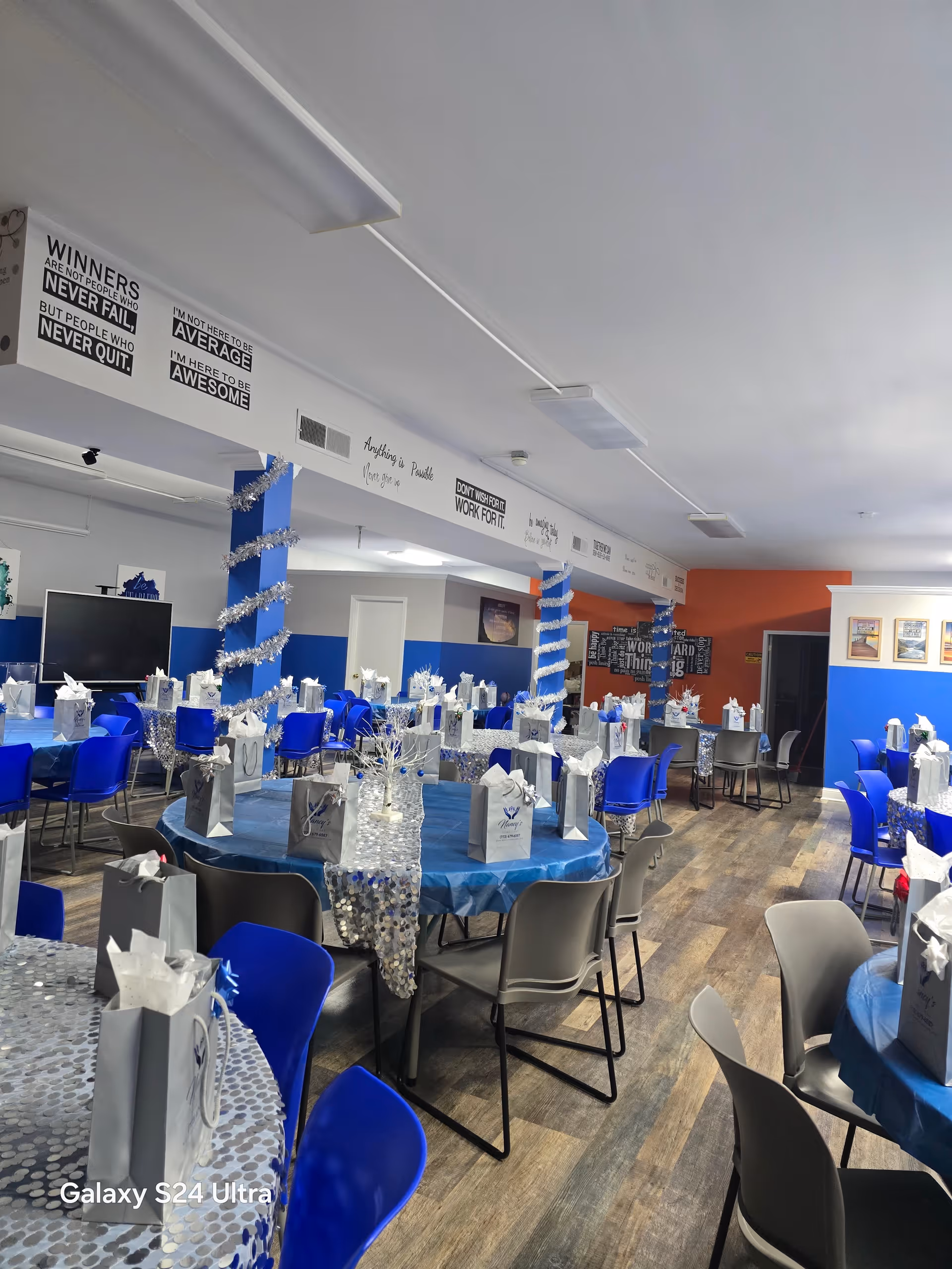 Indoor dining hall with round tables, blue chairs, gift bags, and silver decorations.