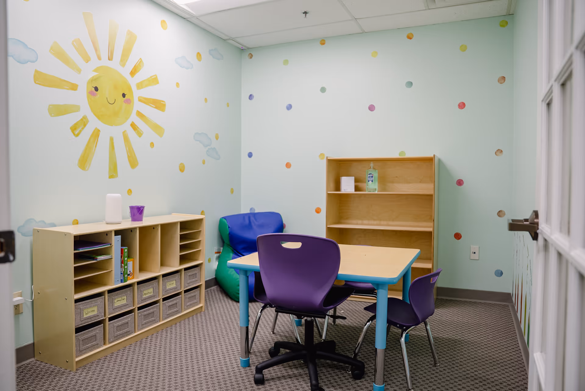 Bright activity room with sun mural, pastel walls, shelves, table, and purple chairs.