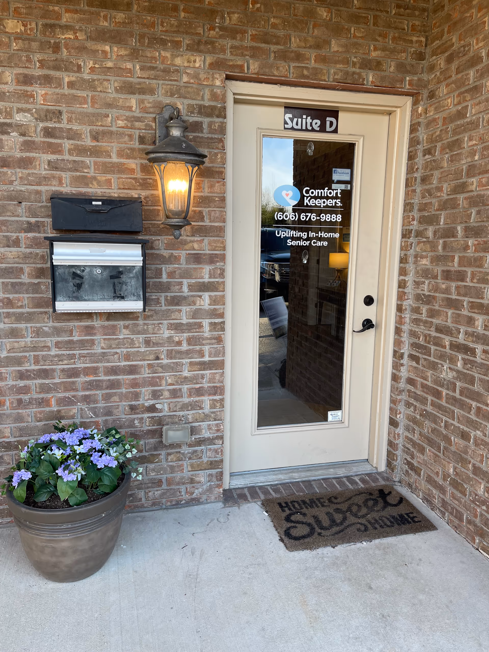 Brick exterior entrance with beige door marked Suite D, lamp, mailbox, and potted flowers.