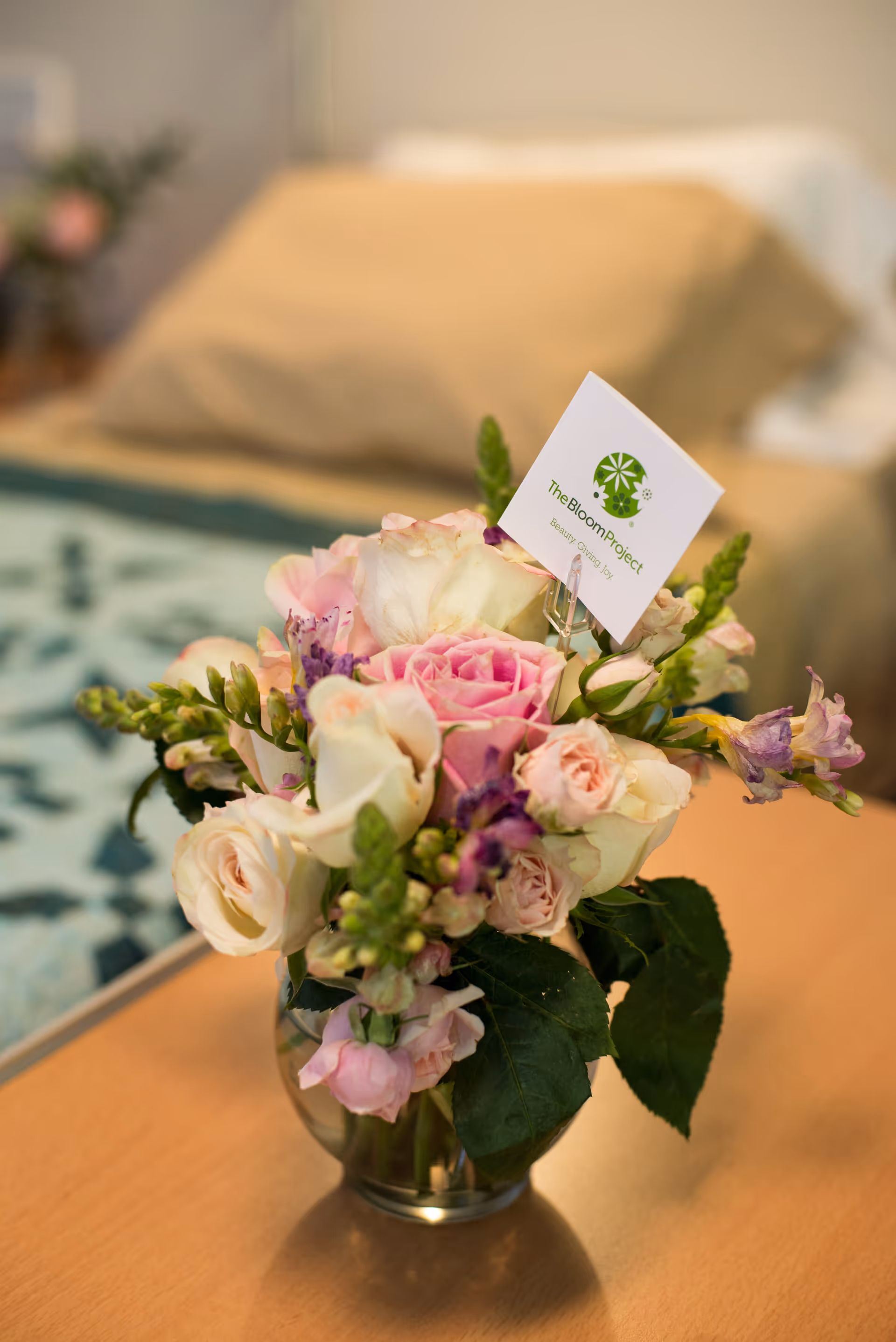A vase of pink and white flowers on a wooden table beside a bed.