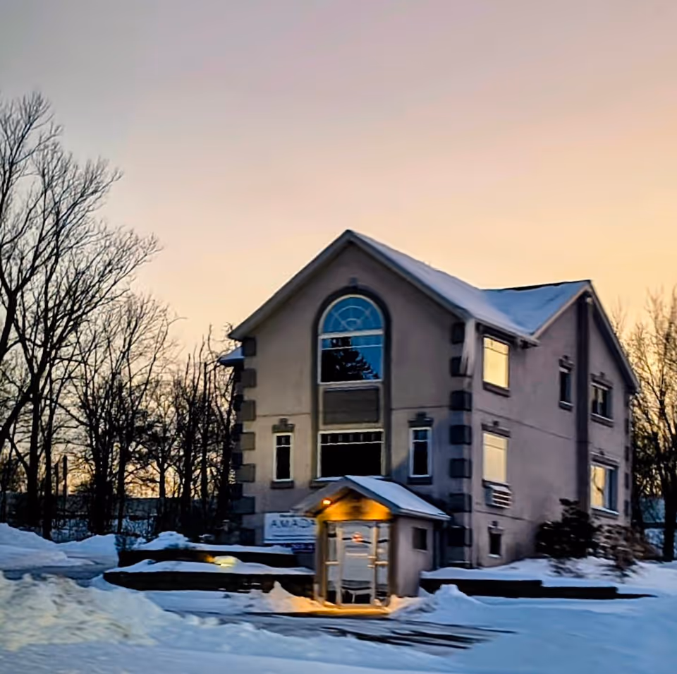 Two-story beige senior living facility with a lit entrance canopy, surrounded by snowy ground.