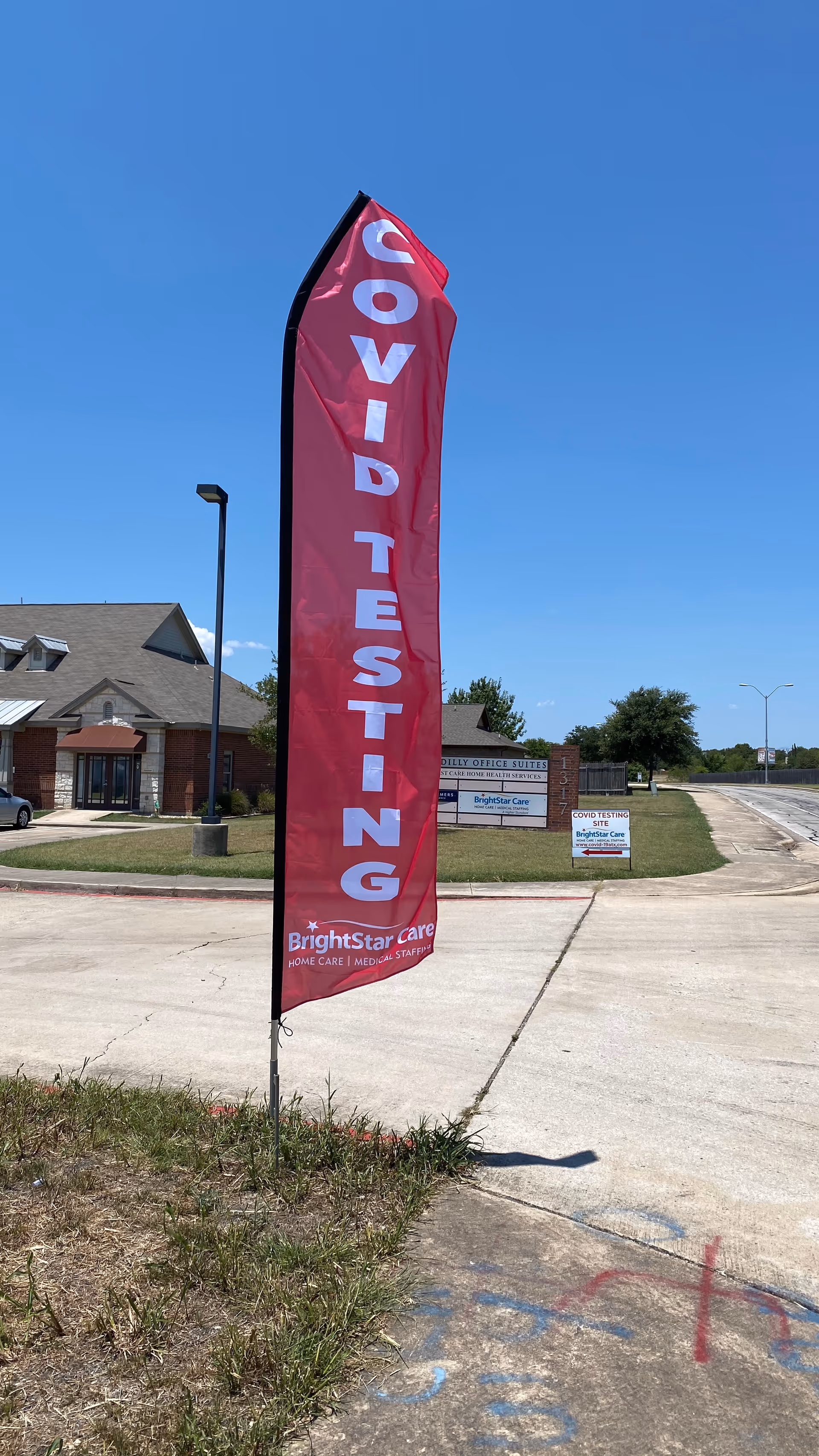 Tall pink COVID testing flag along sidewalk outside BrightStar Care facility under clear blue sky.