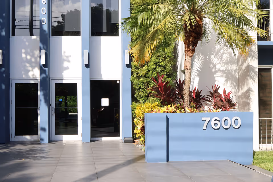 Modern facility entrance with glass doors, palm trees, and a blue 7600 sign.