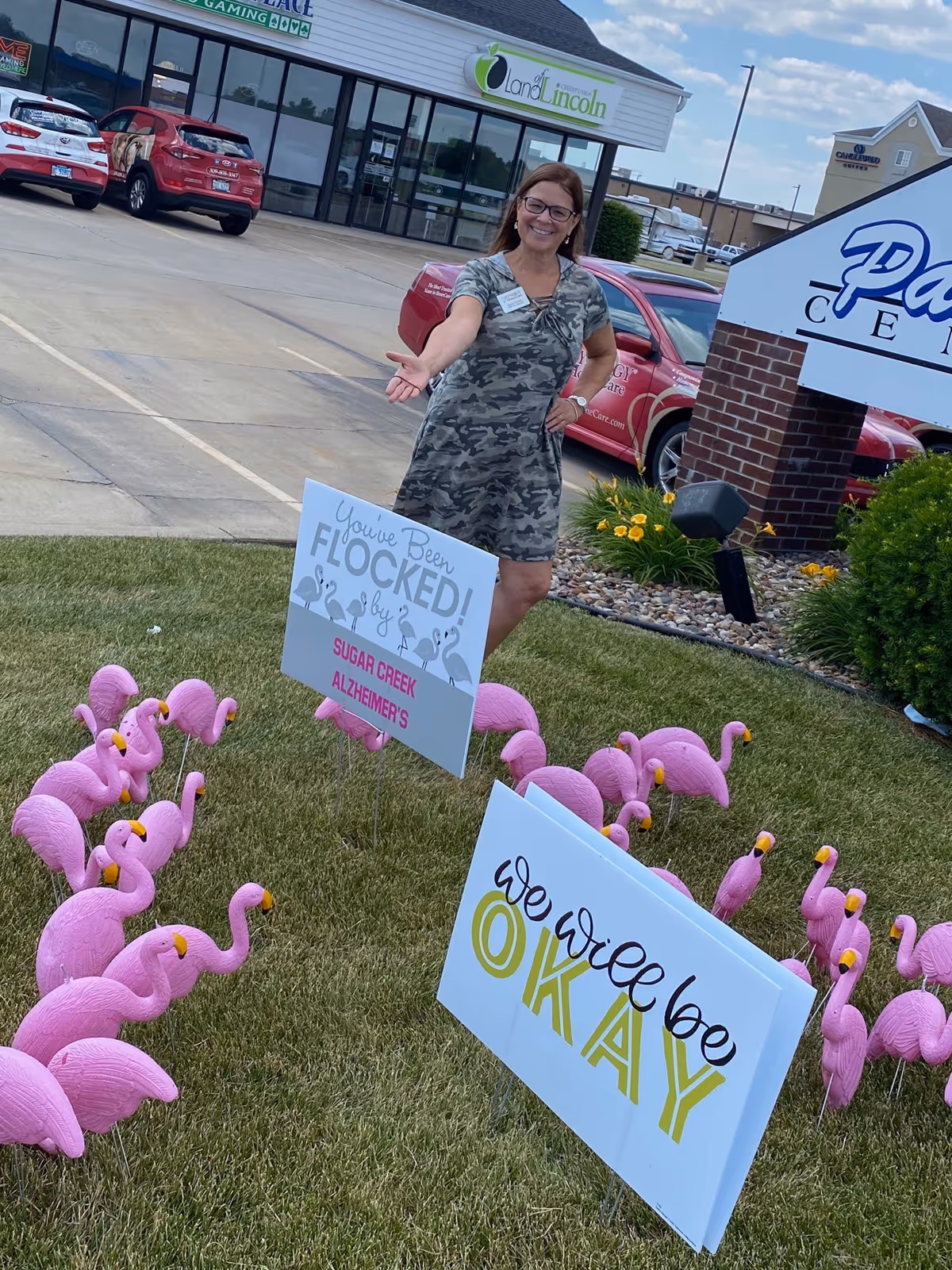 Smiling woman in camo dress stands outside a building, surrounded by pink flamingo decorations and signs.