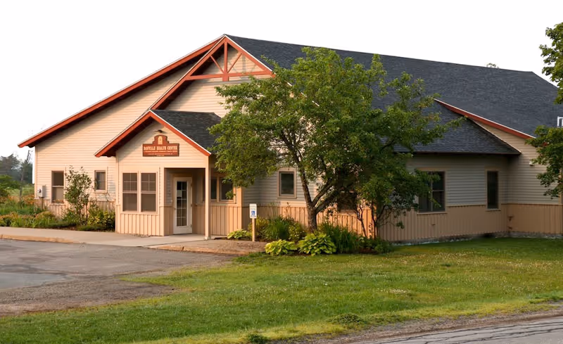 Front view of a beige healthcare facility with trees and parking.