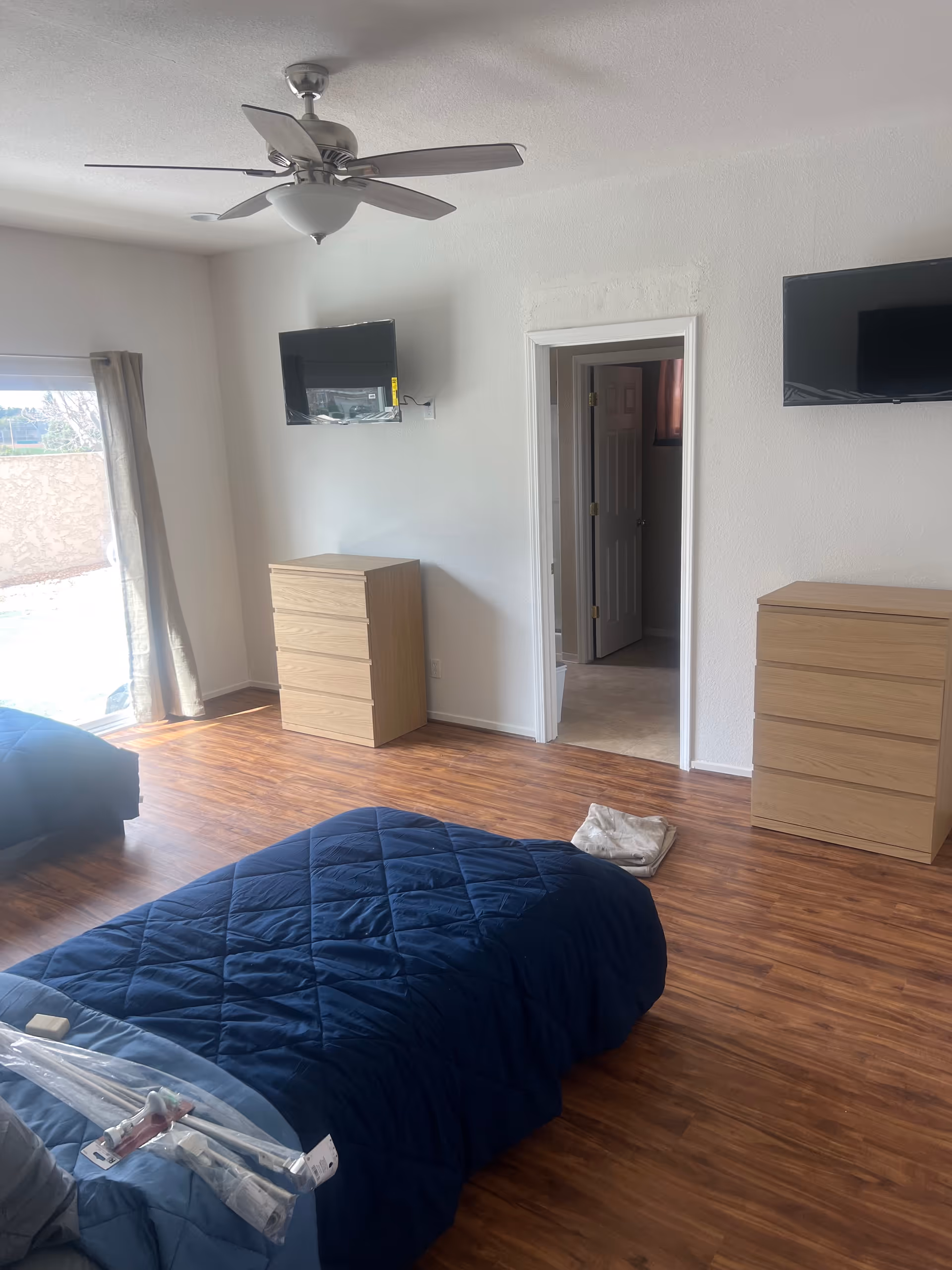 A bedroom with two dressers, wall-mounted TVs, a ceiling fan, and a sliding door to outside.
