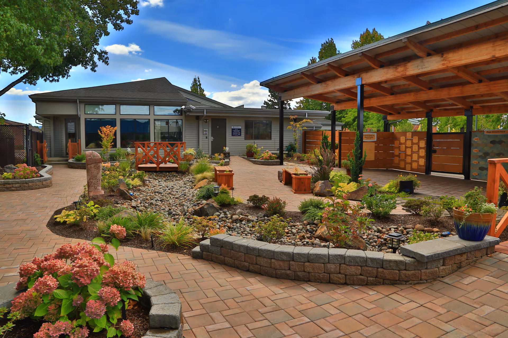 Sunny landscaped outdoor courtyard with brick paths, planters, and a covered seating area.