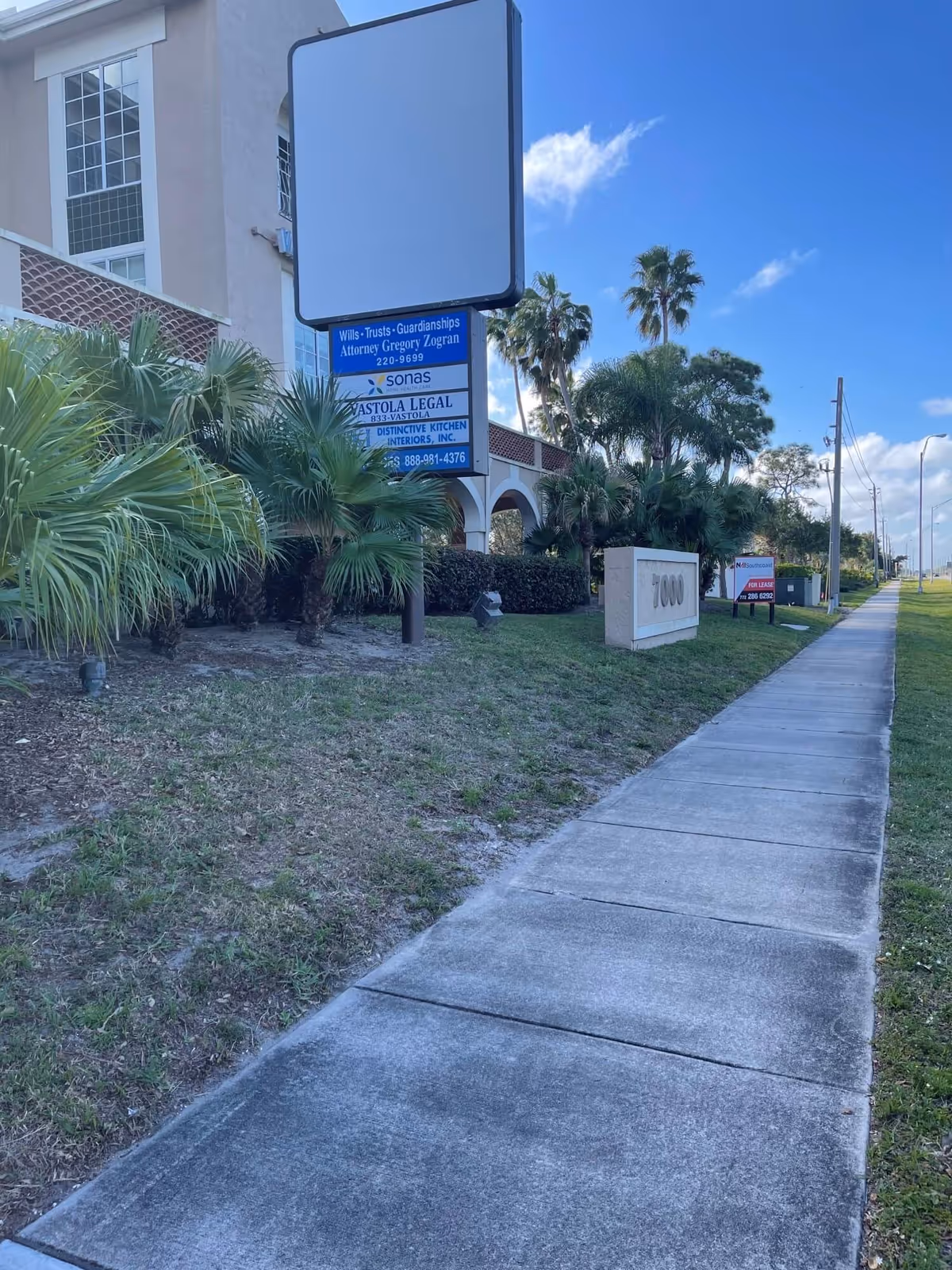 Sidewalk along a facility exterior with palm plants and a large blank sign.
