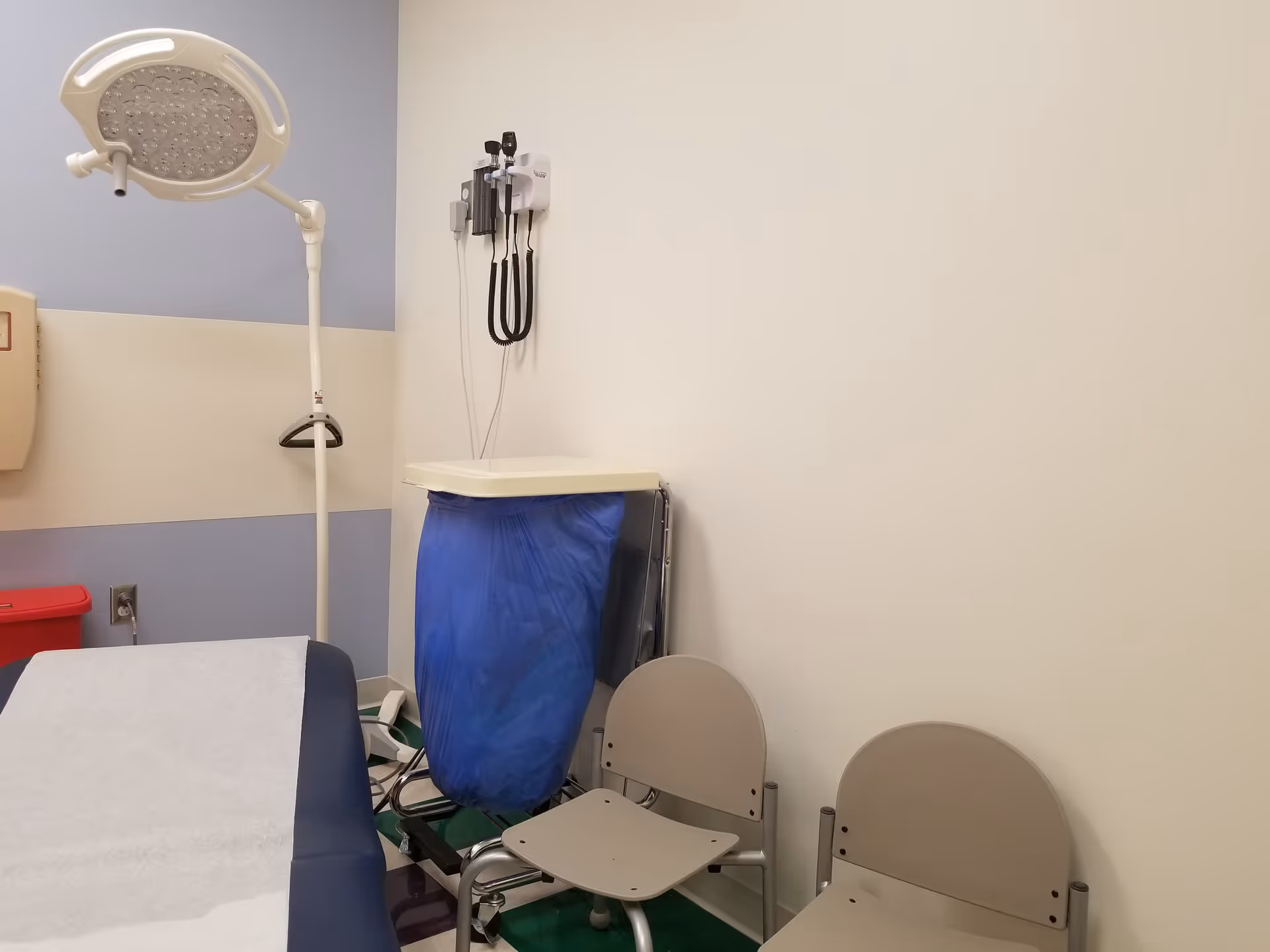 Medical examination room with overhead light, wall-mounted stethoscope, blue waste bag, and two beige chairs.