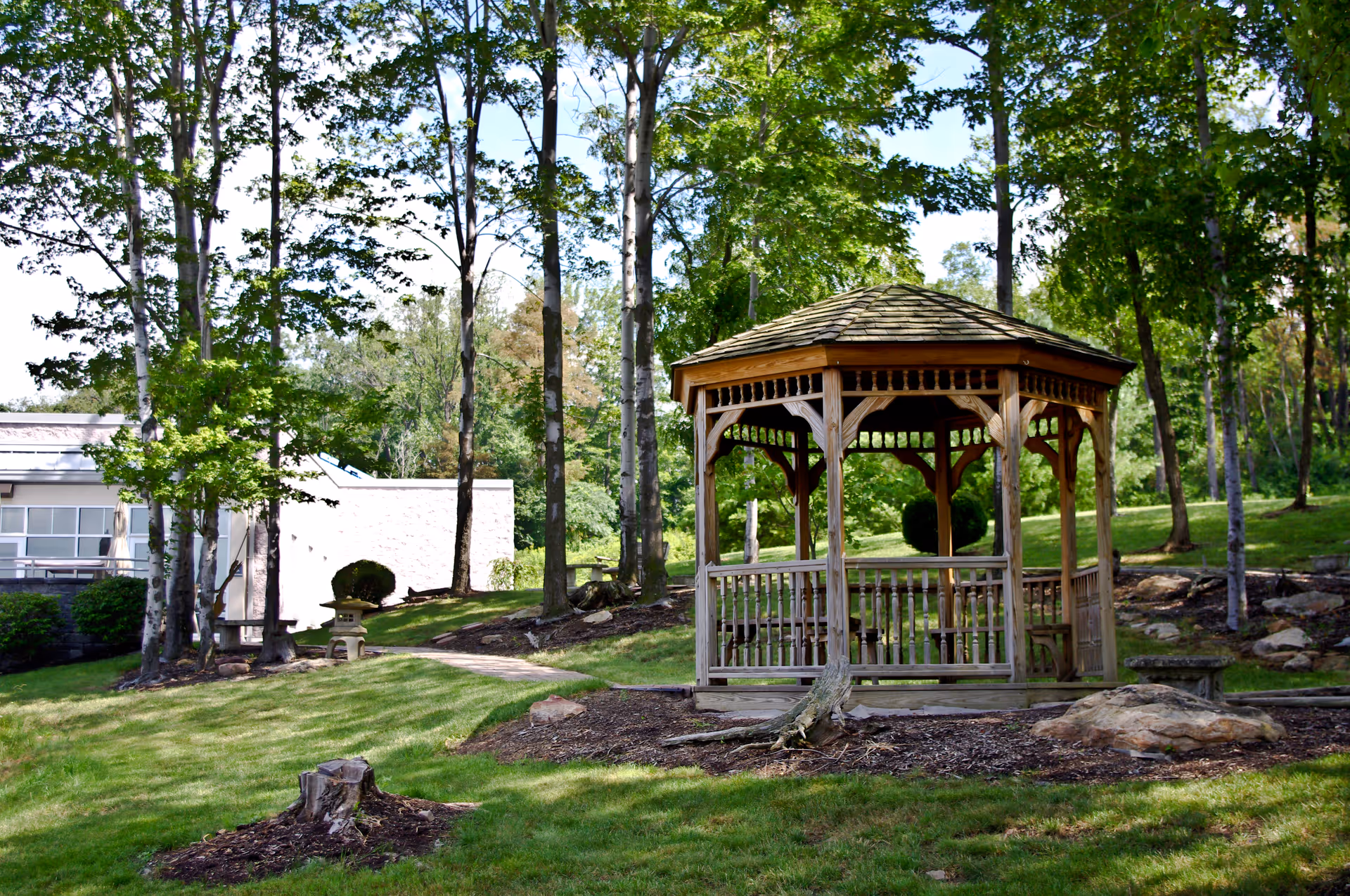Wooden gazebo on a grassy lawn surrounded by tall trees near a building