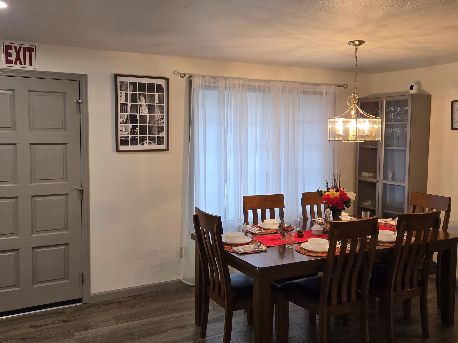 Interior dining room with a wooden table, chairs, chandelier, and white sheer curtains.
