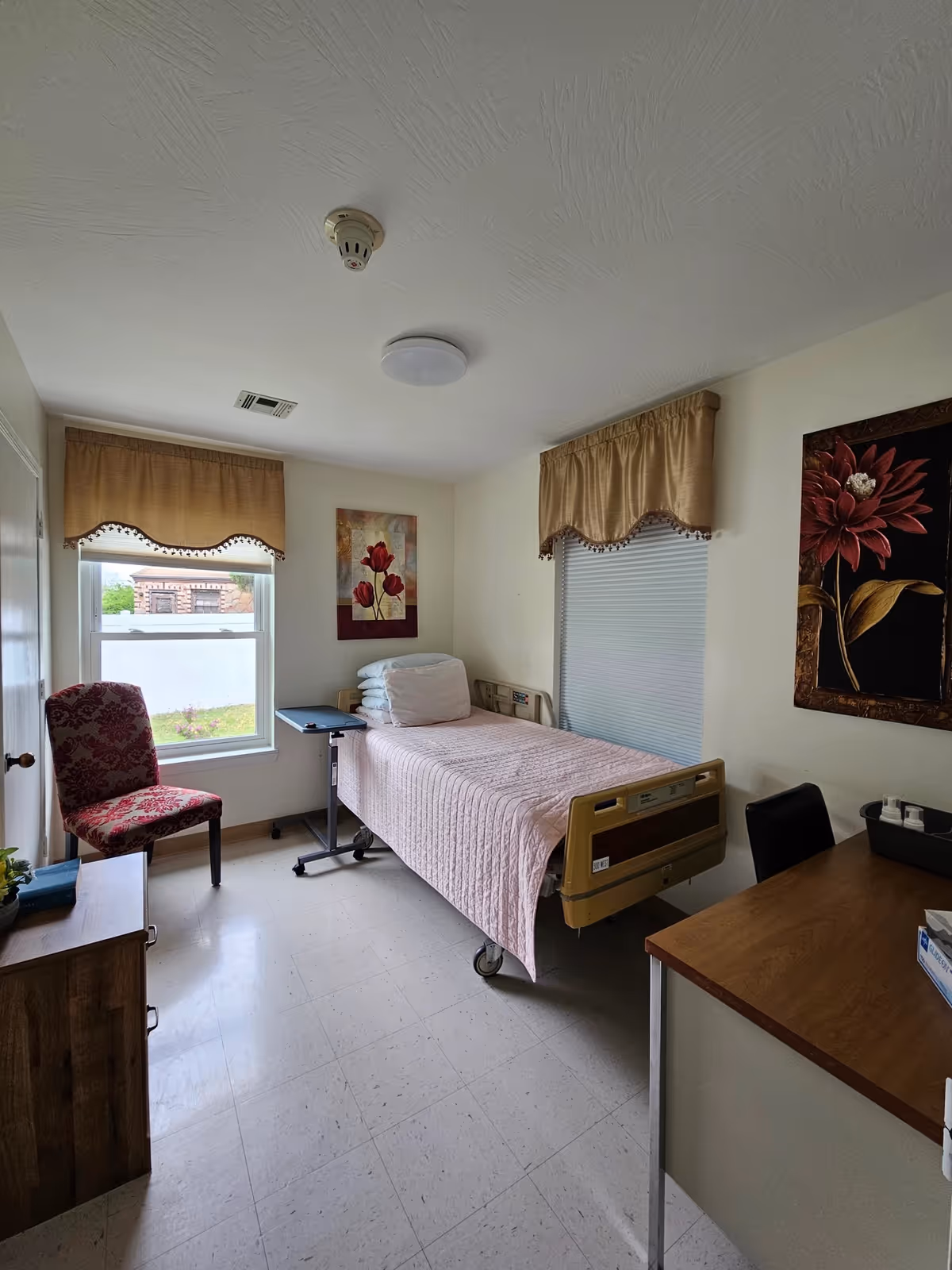 A small patient bedroom with a hospital-style bed, chair, desk, and floral wall art.