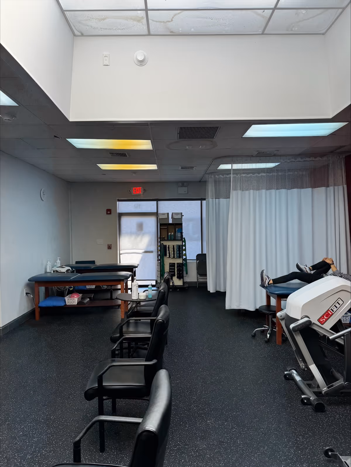 A clinical therapy room with treatment tables, chairs, and a person resting on a bed.
