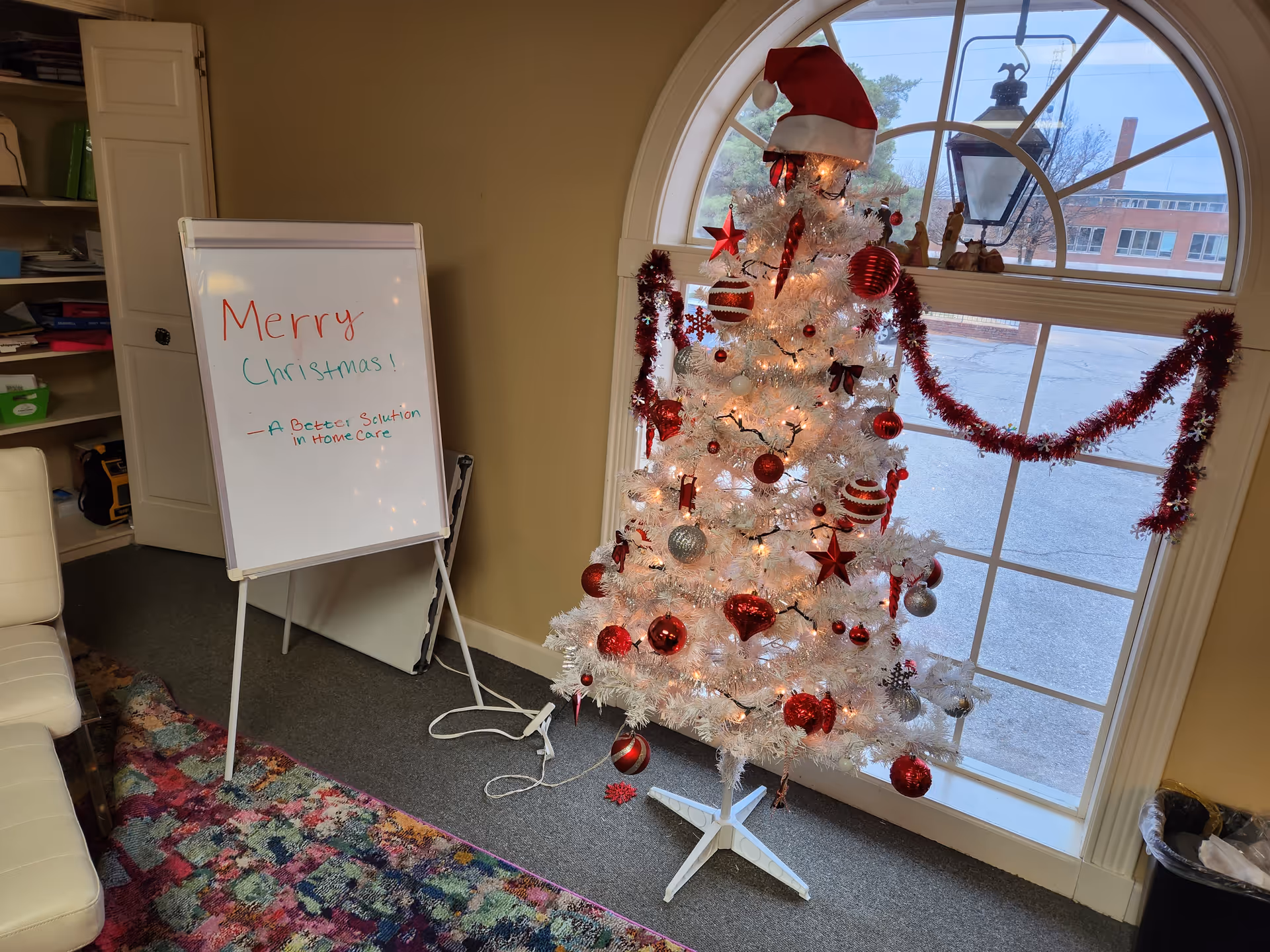 White Christmas tree with red ornaments beside a window, near a whiteboard reading Merry Christmas.