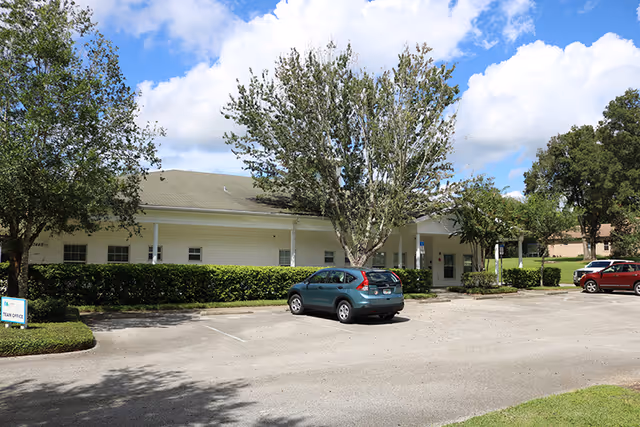 Exterior view of a single-story facility with a parking lot, trees, and blue sky.