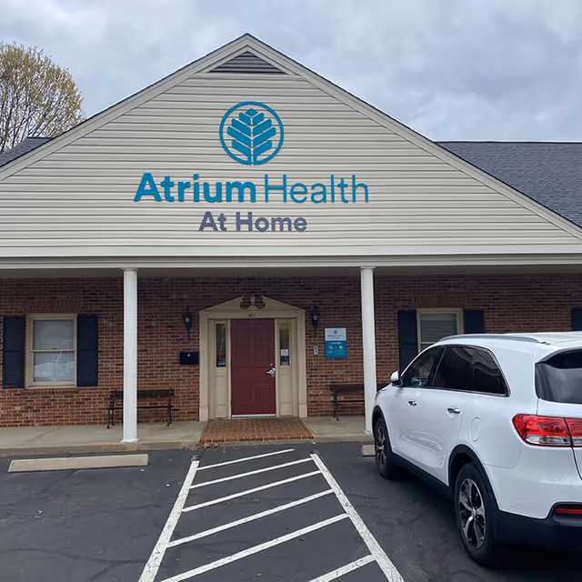 Front exterior of Atrium Health At Home building with red entry door and parked white SUV.