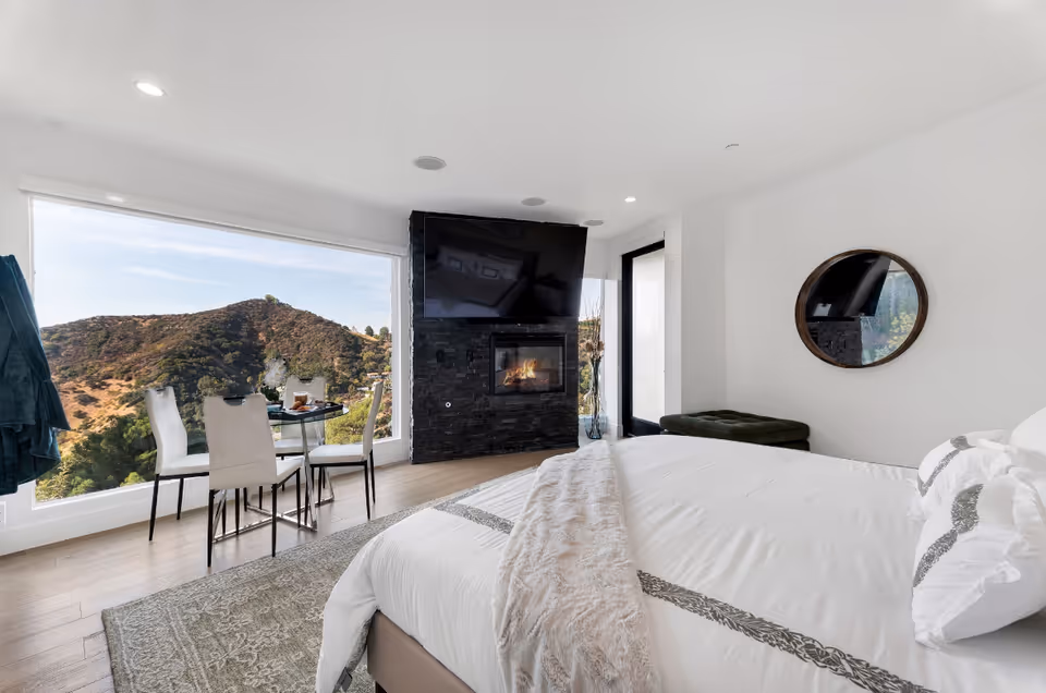 Bright bedroom with a wall-mounted TV above a fireplace and a glass table by the window.