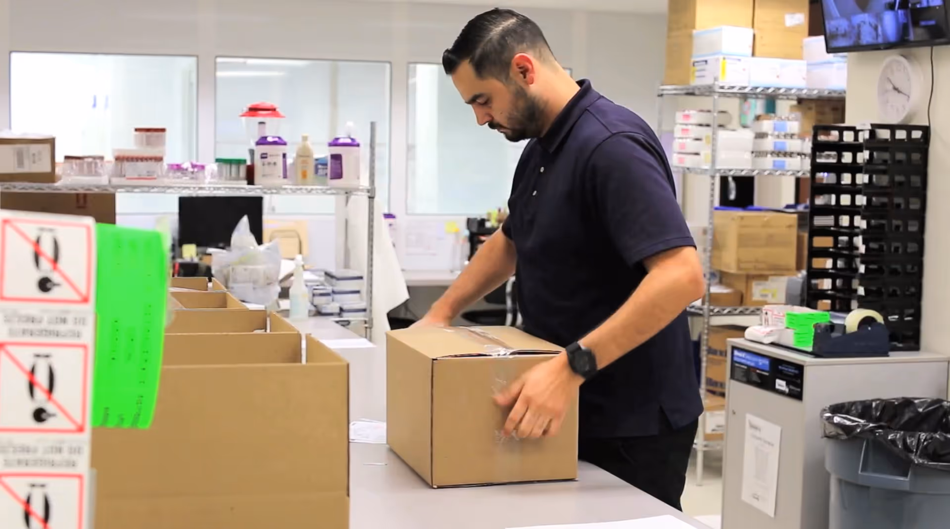 A KabaFusion staff member seals a cardboard box in a cluttered medical supply room.
