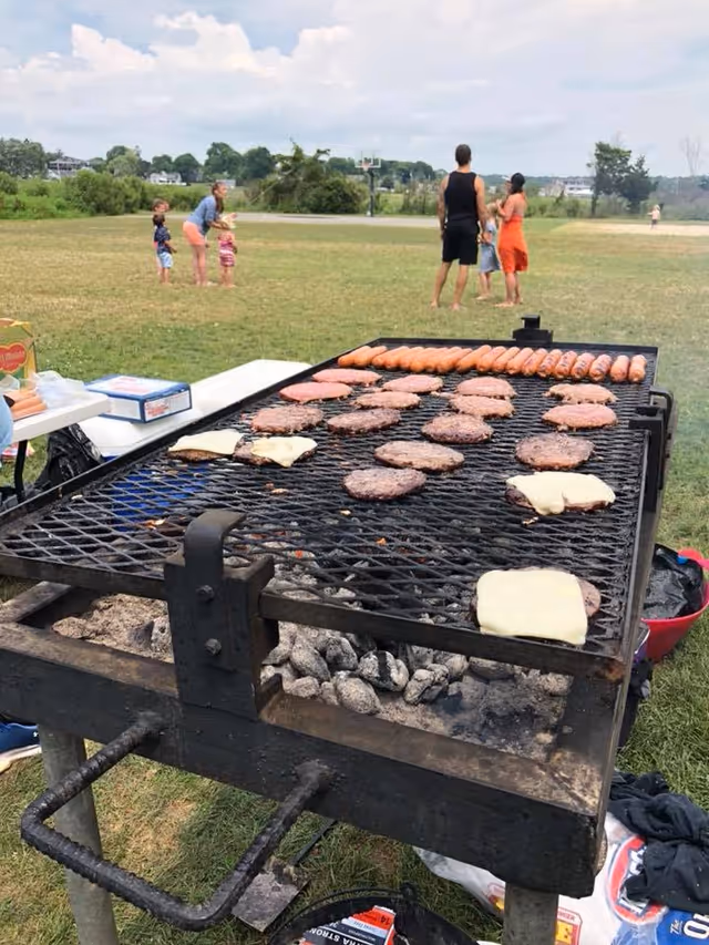 Outdoor barbecue grill with burgers and cheese on a large grill; people nearby in a park