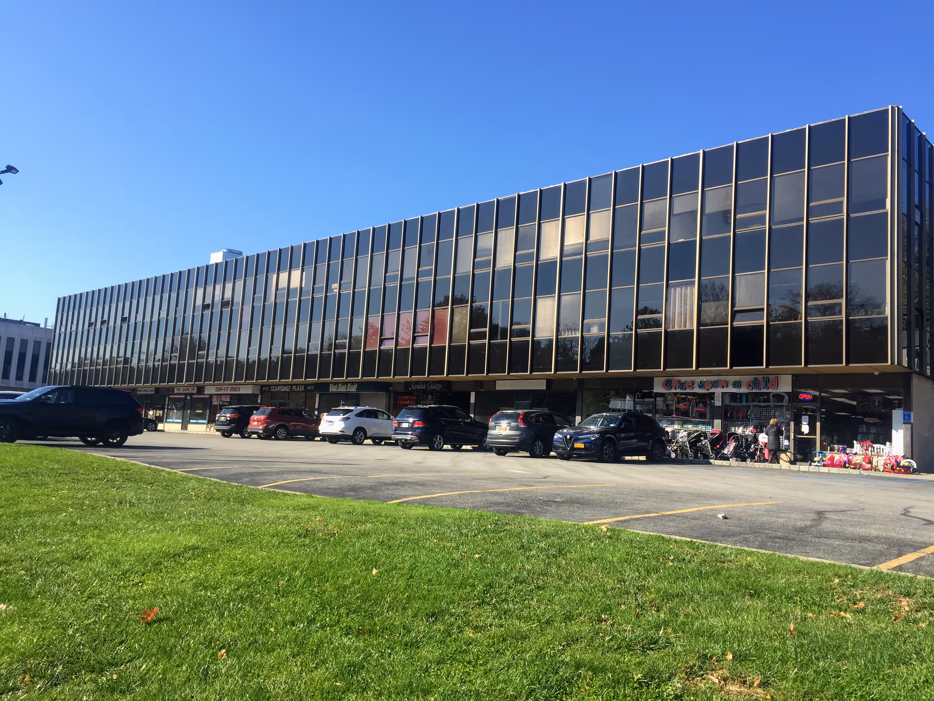 Two-story glass-front building with storefronts, parking, and a grassy foreground.