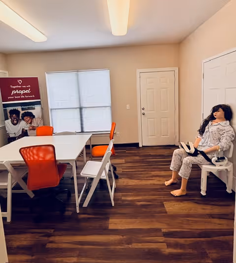 A training room with white tables, orange chairs, and a lifelike mannequin seated by the door.
