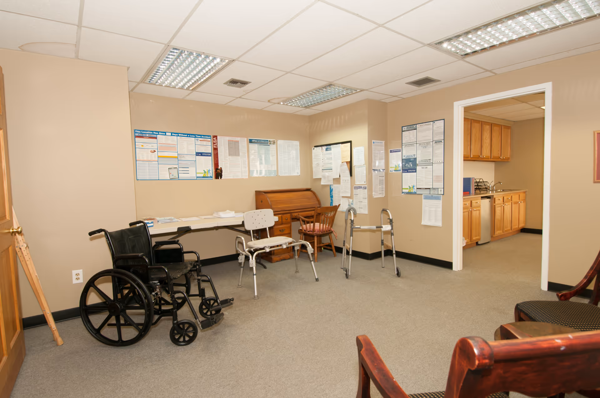 Interior facility lounge with wheelchairs, walkers, chairs, and a kitchenette in background.