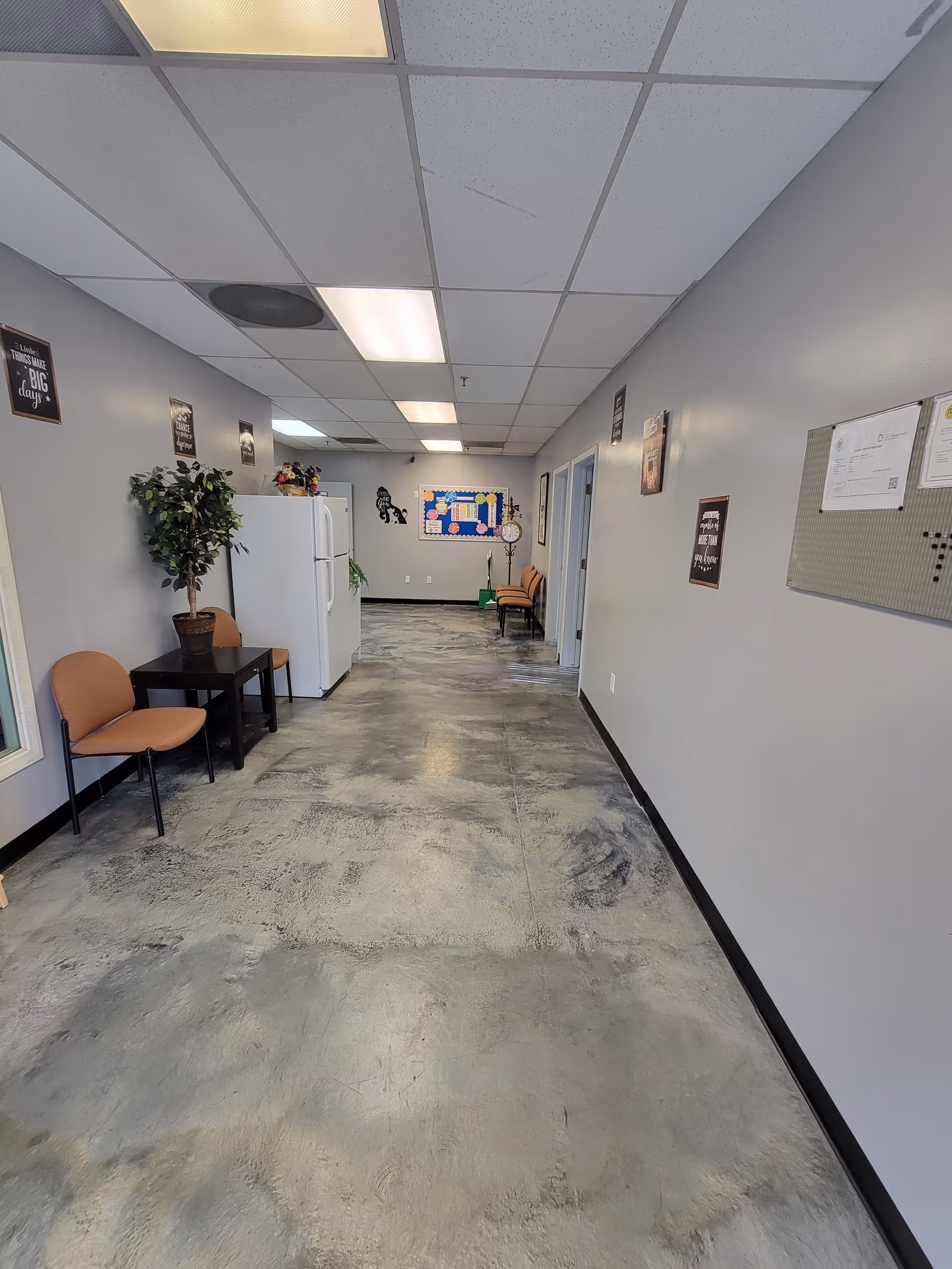 Wide hallway in a senior living facility with chairs, a plant, and bulletin boards along walls.